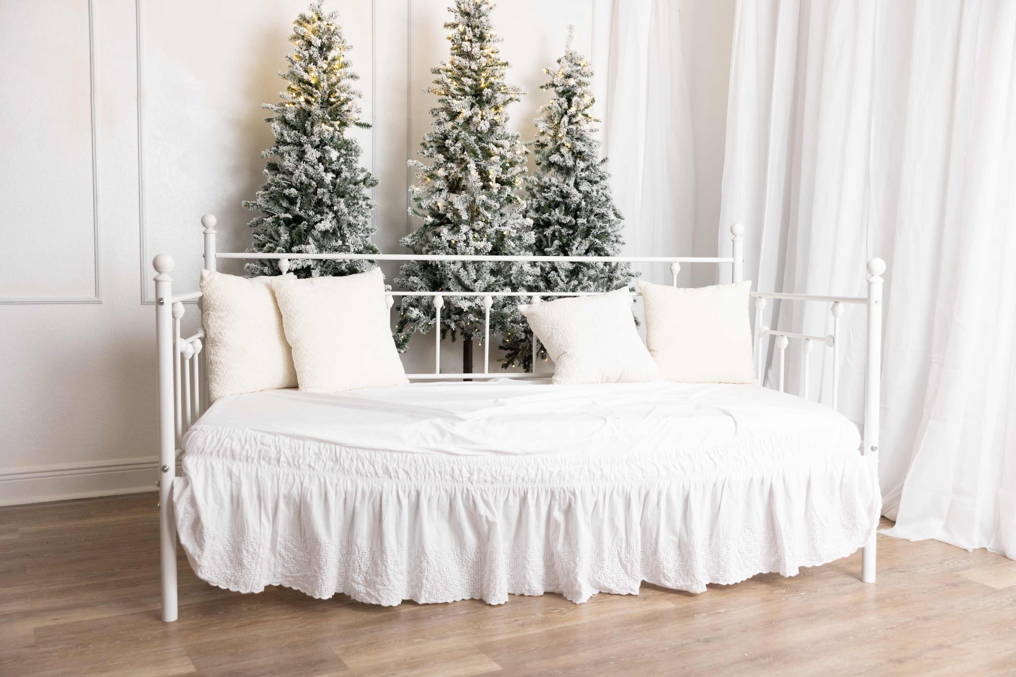 A white daybed with pillows is positioned in front of two decorated, snowy Christmas trees.
