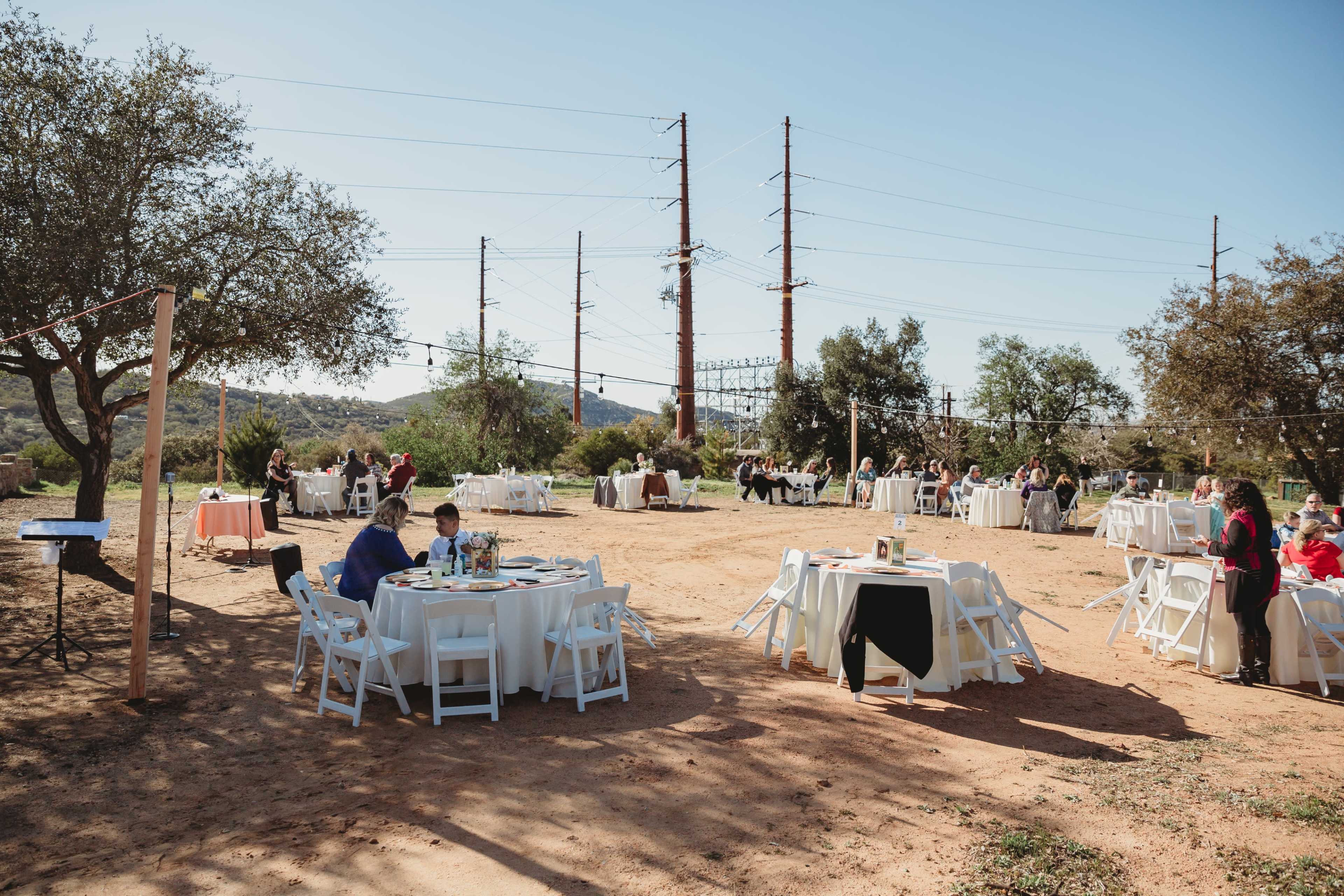 A reception area with several round tables set up outdoors, surrounded by trees and power lines, hosts guests enjoying a meal.