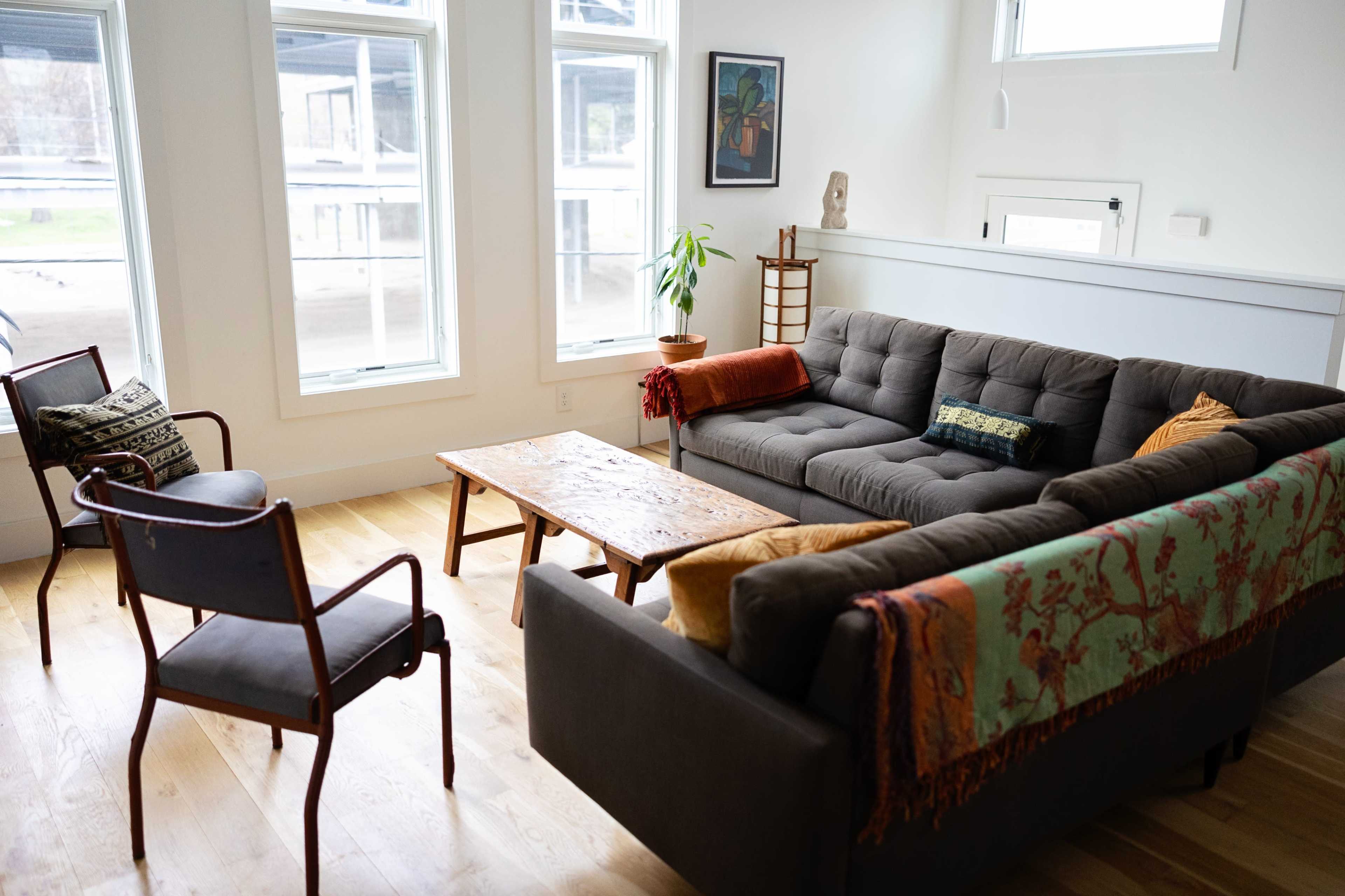 The image shows a modern living room with a gray sofa, two chairs, a wooden coffee table, and large windows allowing natural light.