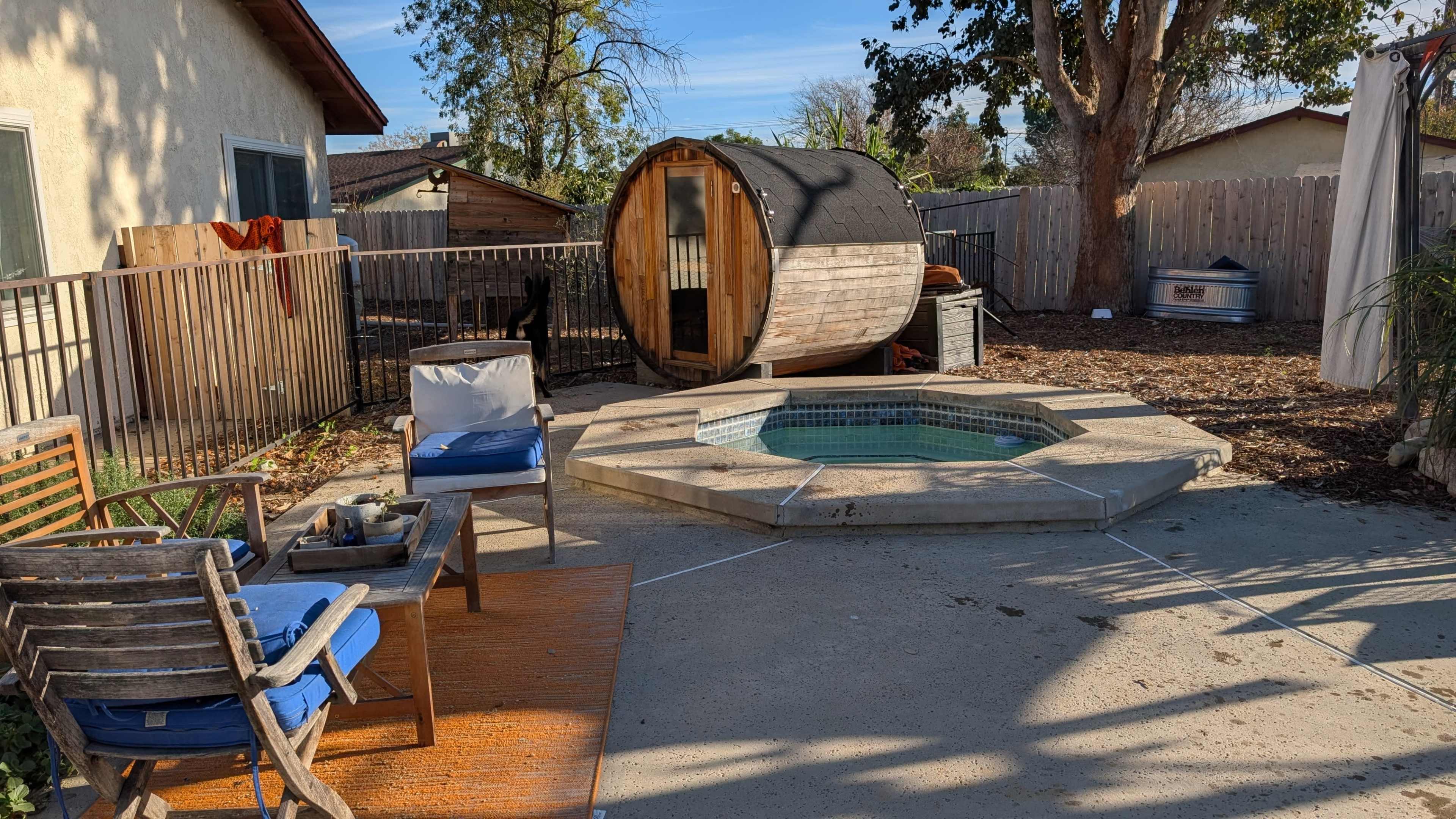 The image shows a backyard with a circular wooden sauna, a small round hot tub, and outdoor seating that includes wooden chairs and a blue cushion on a patio area.