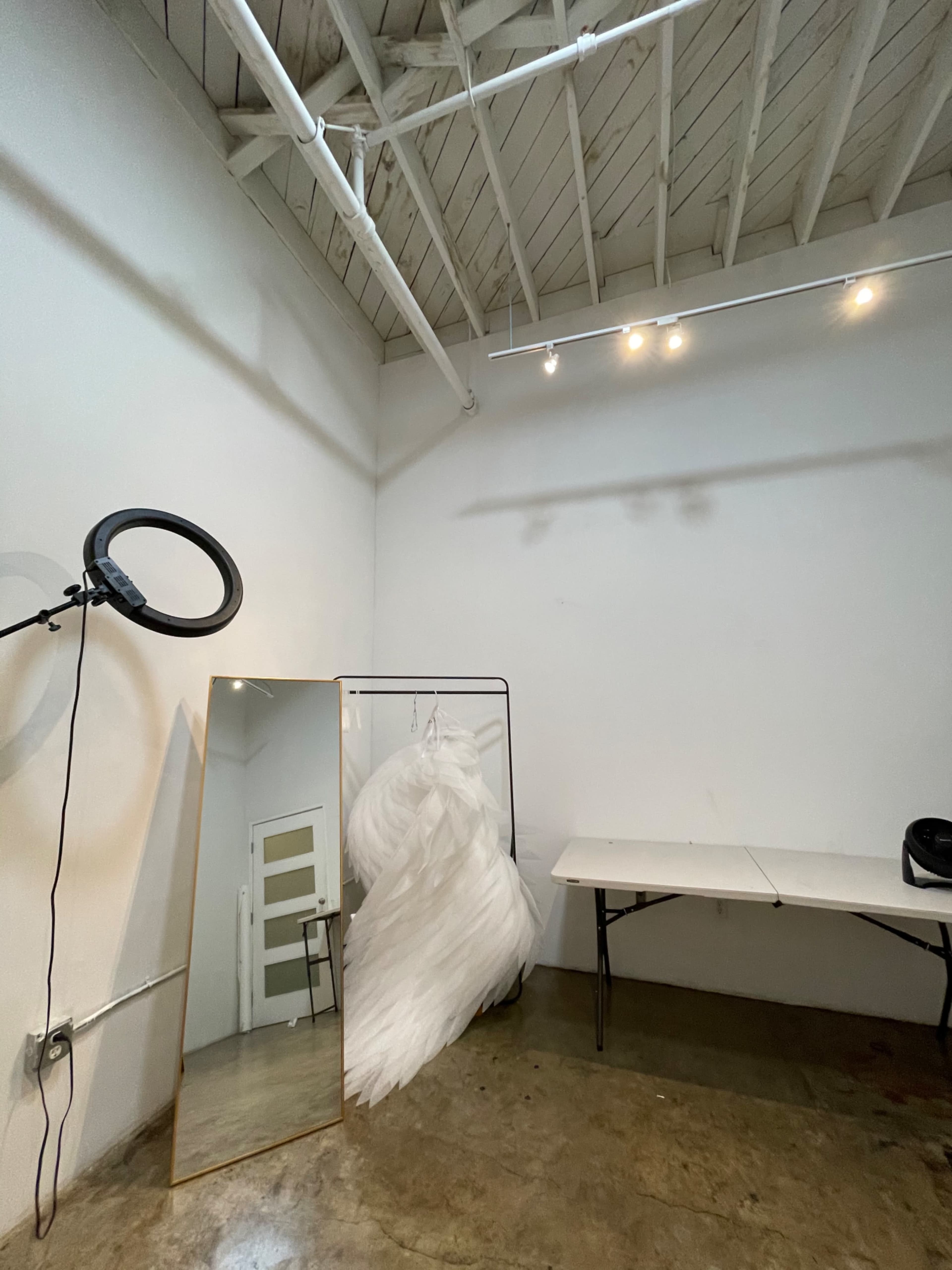 The image depicts a minimalist studio space featuring a large mirror, a clothing rack with a white dress, and a table against a white wall with exposed beams.