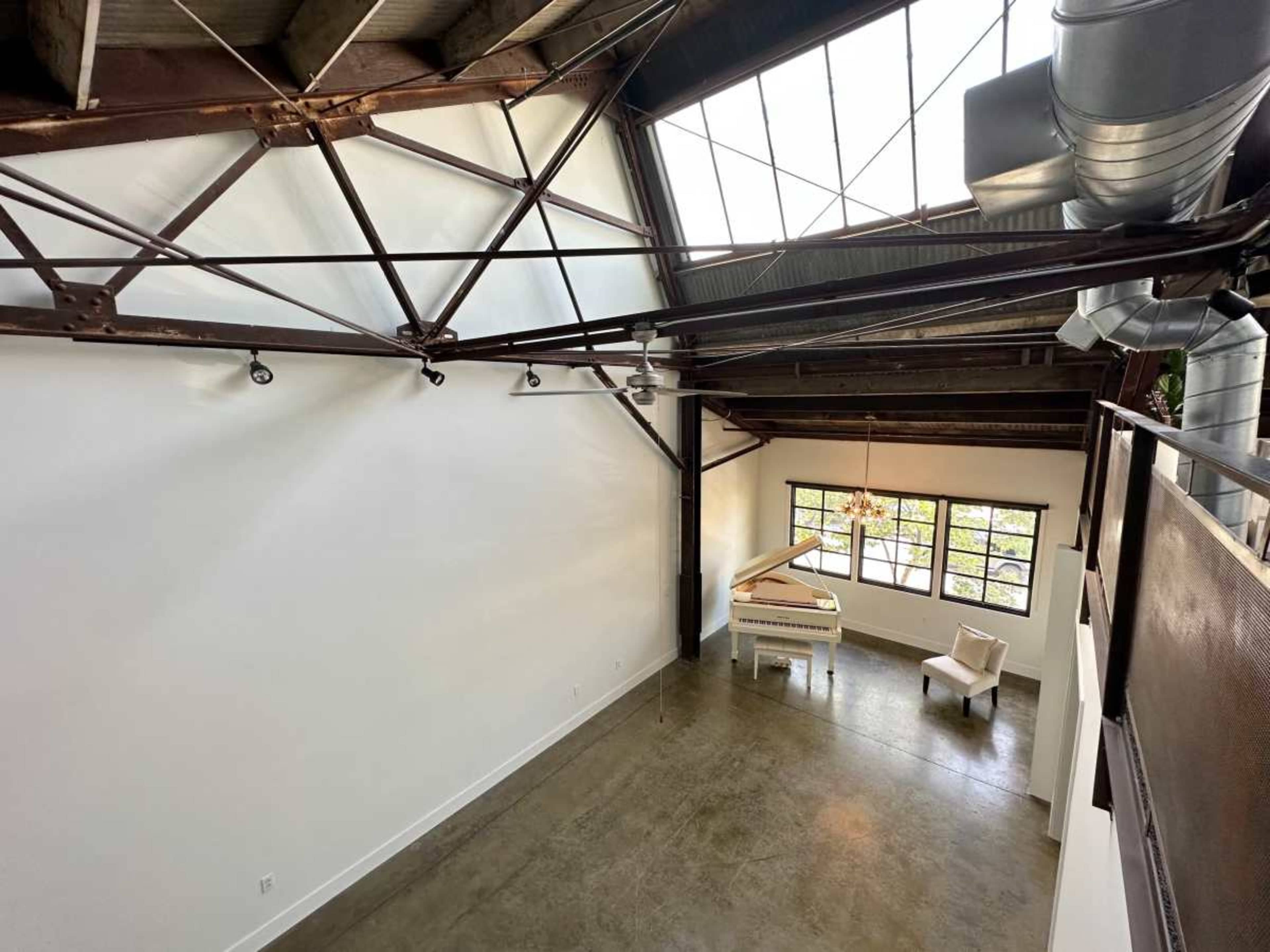 The image shows a spacious, industrial-style interior with high ceilings, exposed metal beams, and large windows allowing natural light to fill the room, which contains a piano and a chair.