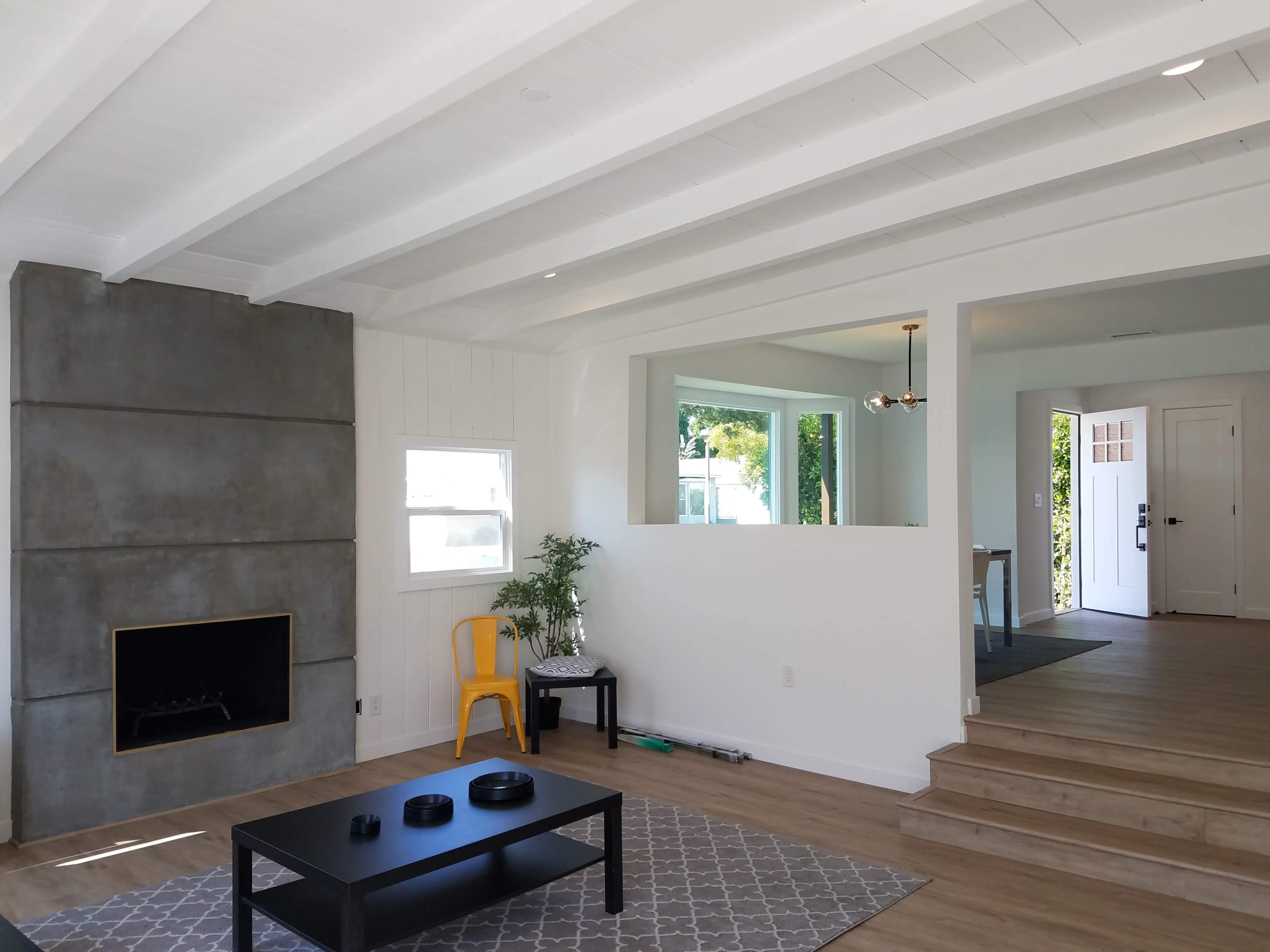 A modern living room with a concrete fireplace, a black coffee table, a gray area rug, and an open layout leading to a staircase and an entryway.