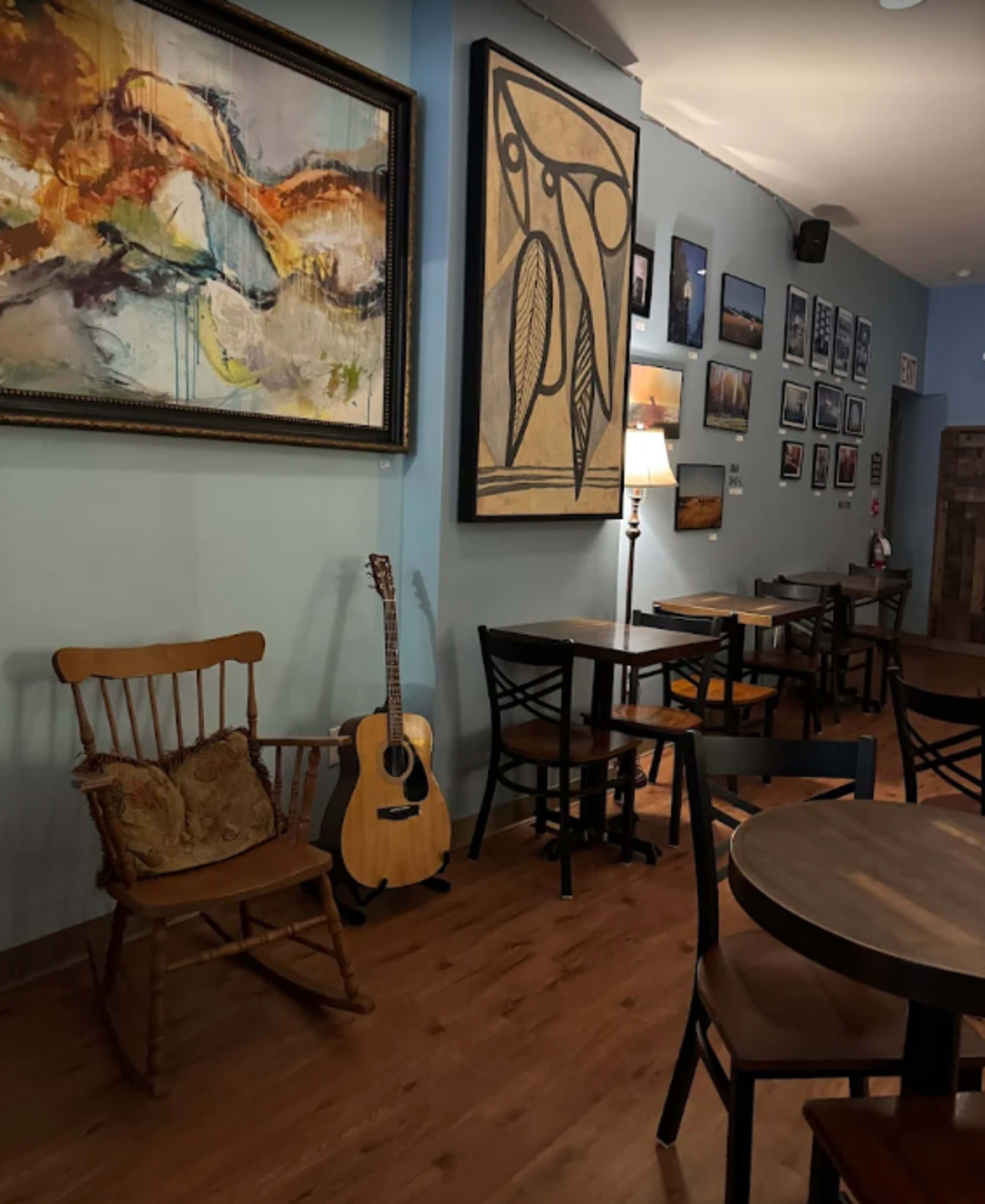 The image shows a cozy café interior with wooden tables, a rocking chair, an acoustic guitar, and various artworks on the walls.