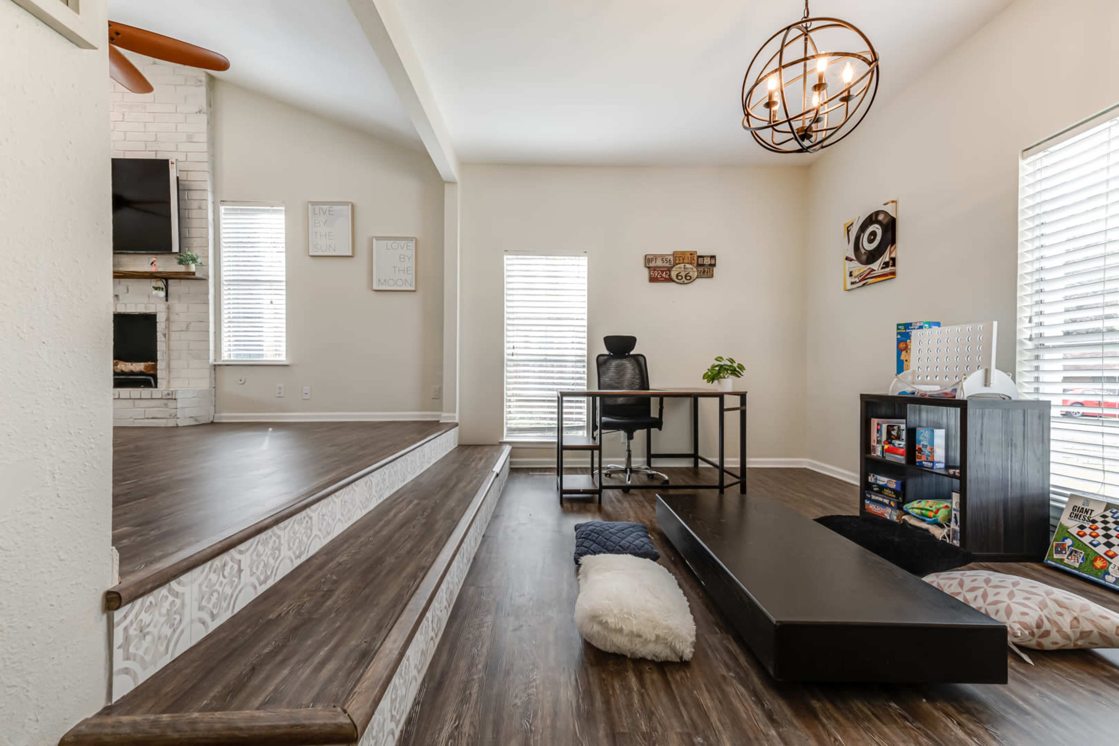 A spacious living area featuring a desk and chair, a large step down to a cozy seating area with cushions, and a fireplace with a television mounted above it.