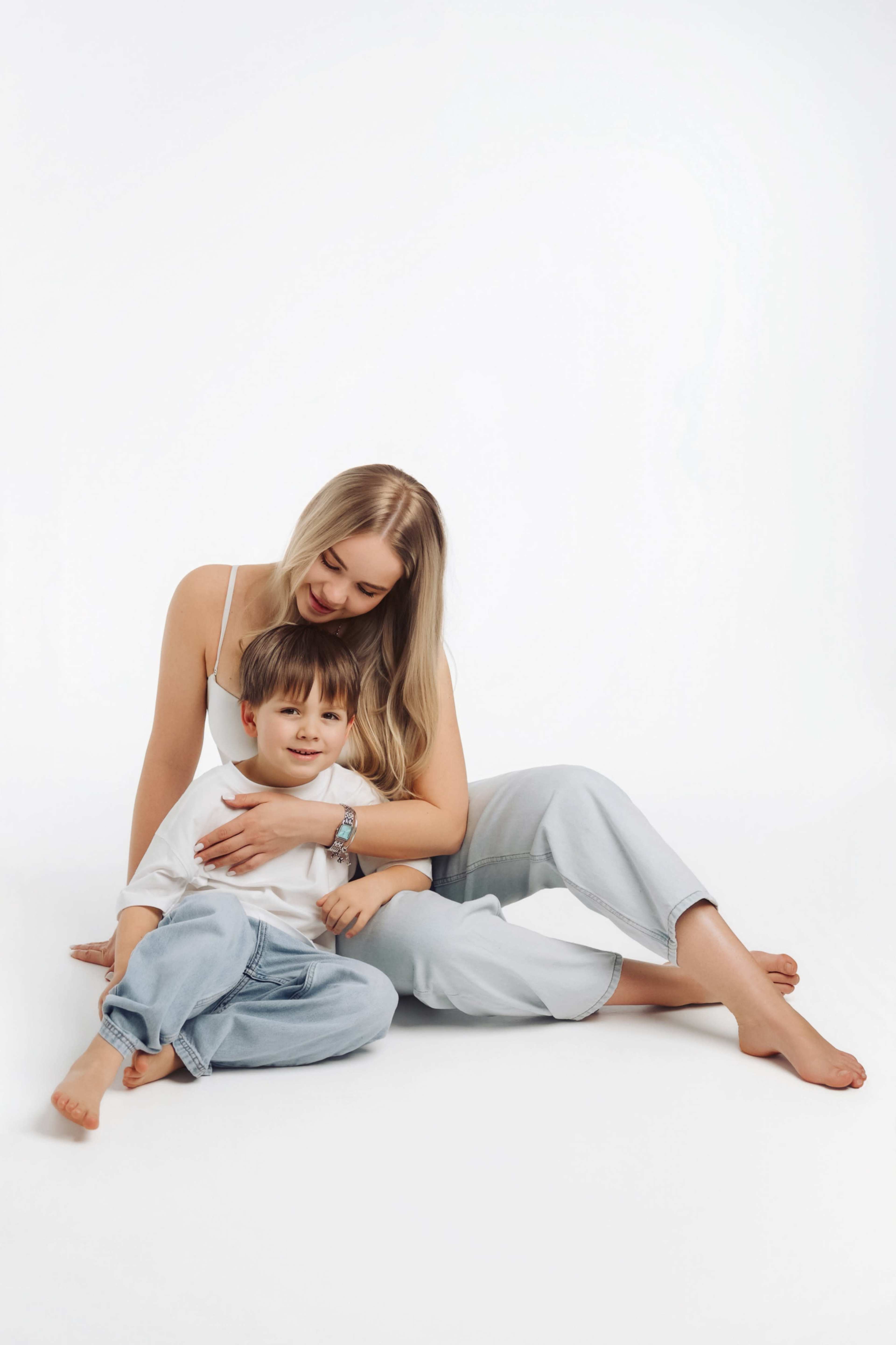 A woman sits on the floor, embracing a young boy who is seated beside her, both wearing casual clothing against a plain white background.
