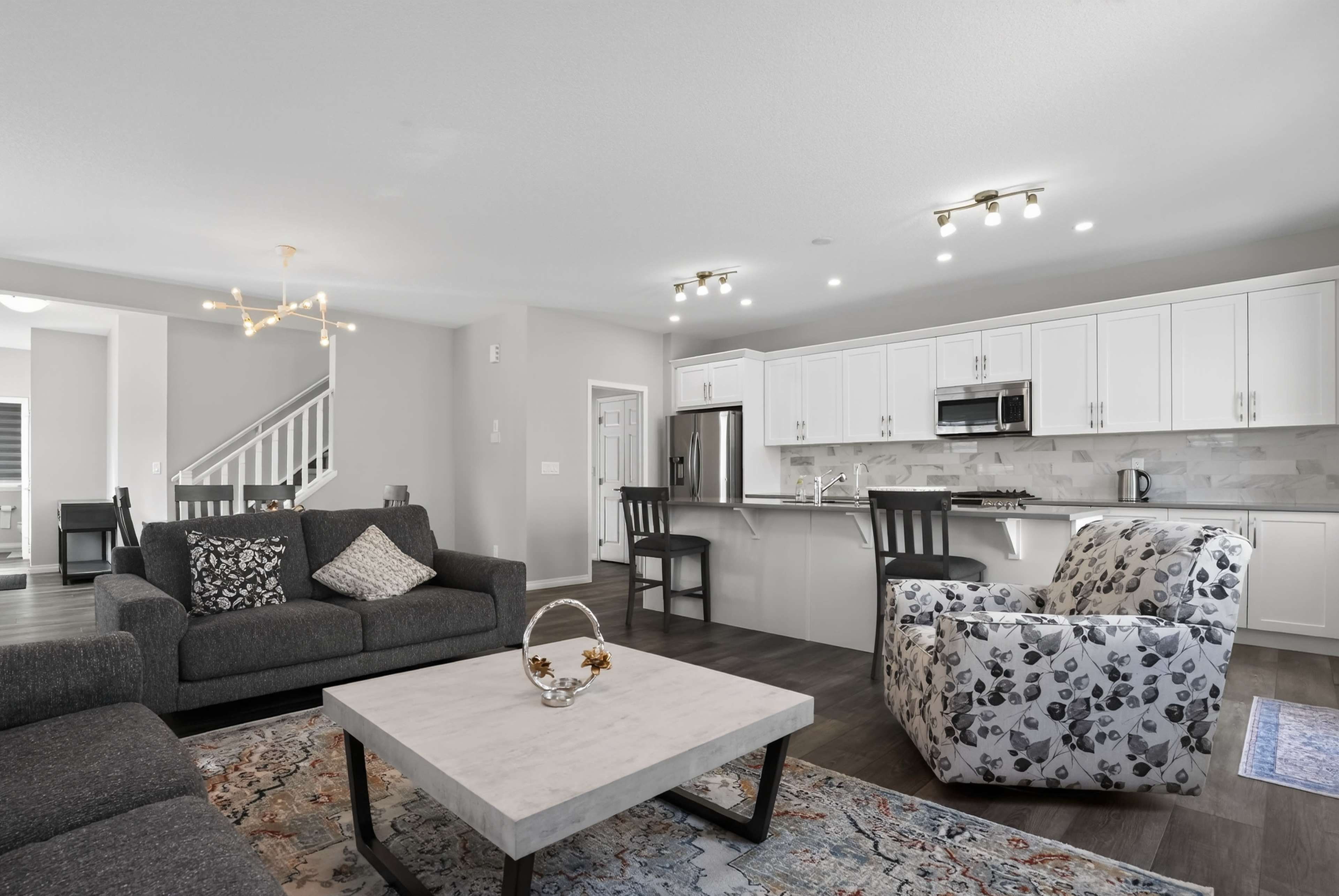 The image shows a modern living room with a gray sofa, an armchair with a floral pattern, and a coffee table in front of a kitchen featuring white cabinetry and stainless steel appliances.