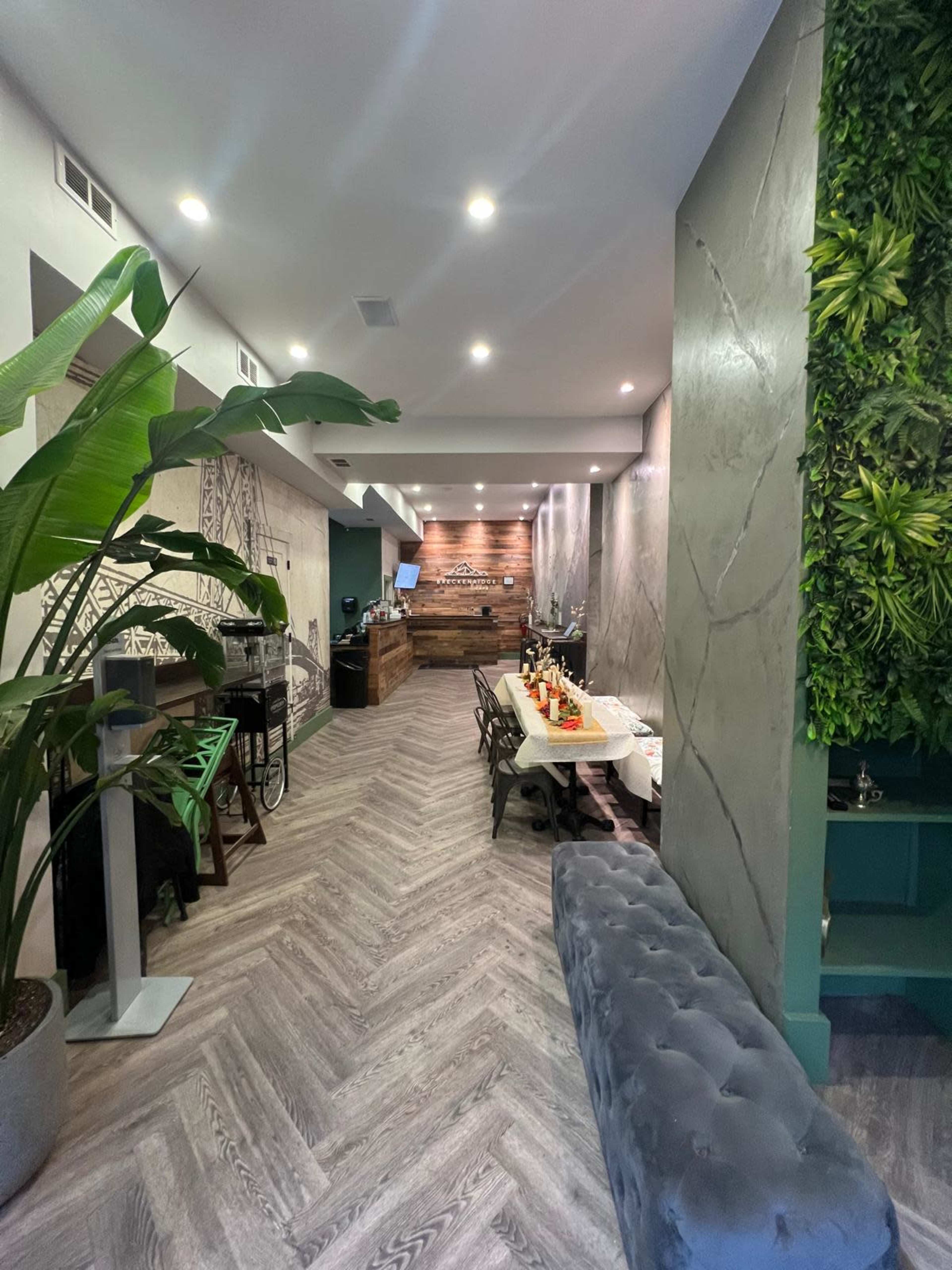 The image shows a modern interior of a café or restaurant with a wooden front desk, a green vertical garden, and herringbone-patterned flooring.