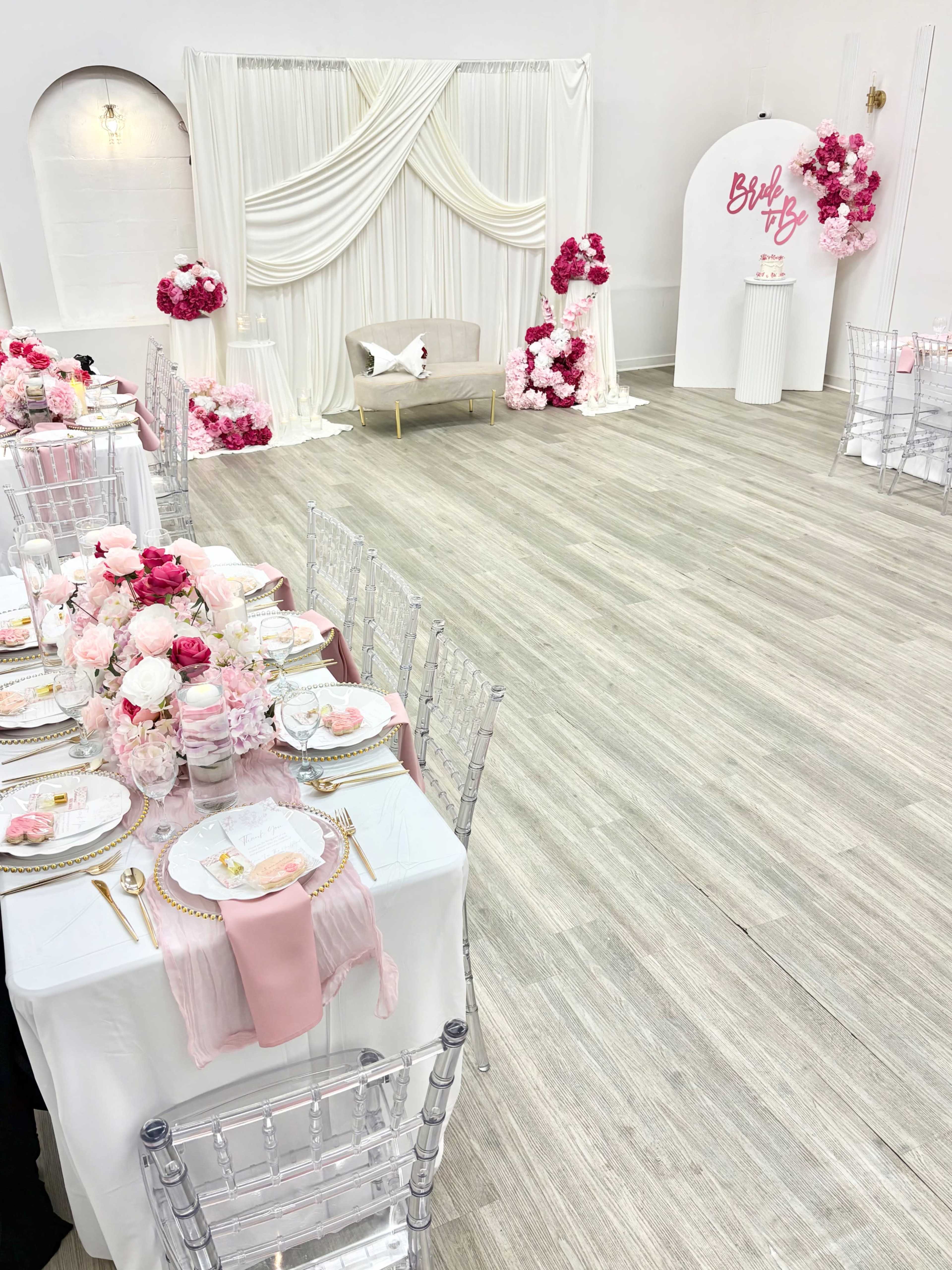 The scene features an elegantly decorated event space with a dining setup, including a white tablecloth, pink floral arrangements, and a backdrop of draped fabric.