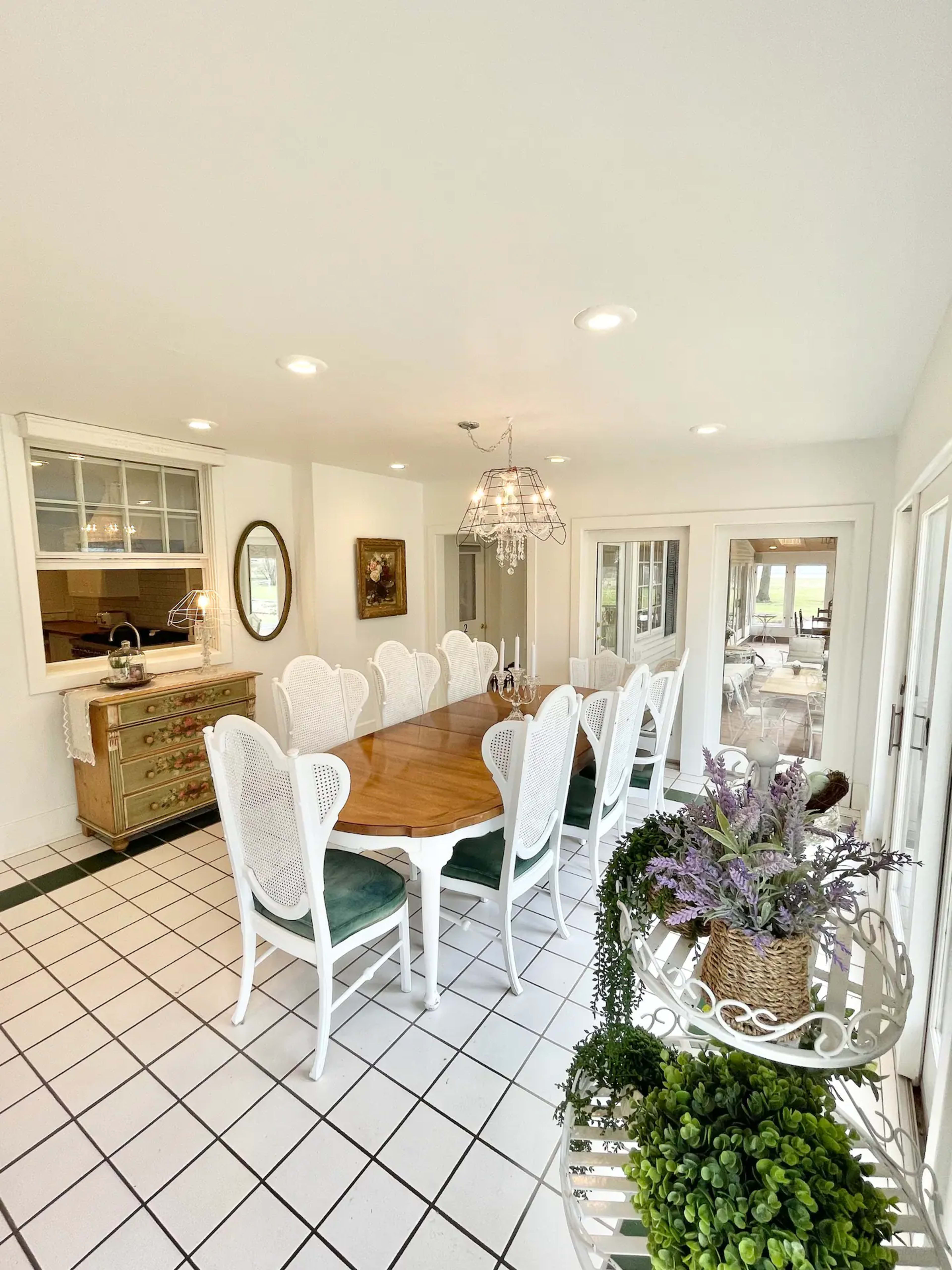A bright dining room featuring a large wooden table surrounded by white chairs, with a sideboard and decor items in the background.