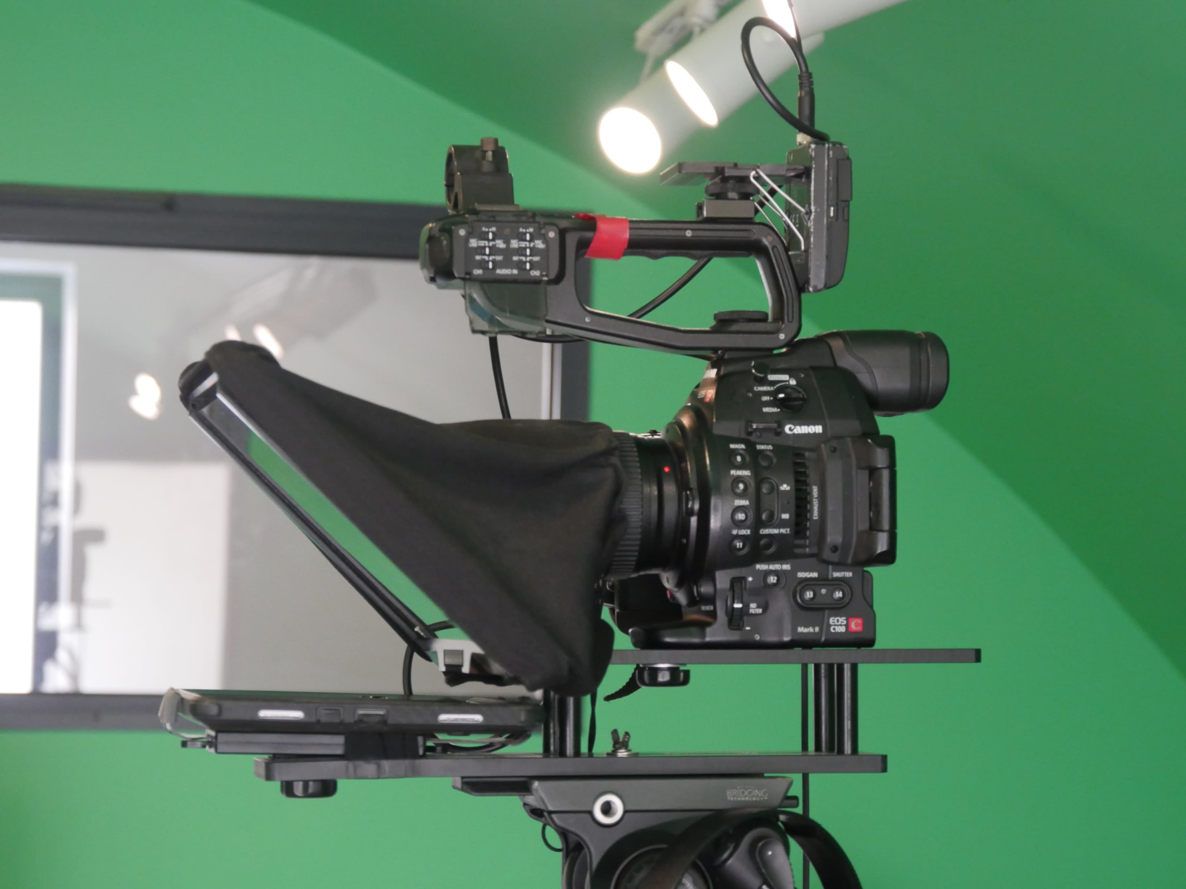 A Canon camera is set up on a tripod with a teleprompter and positioned in front of a green backdrop.