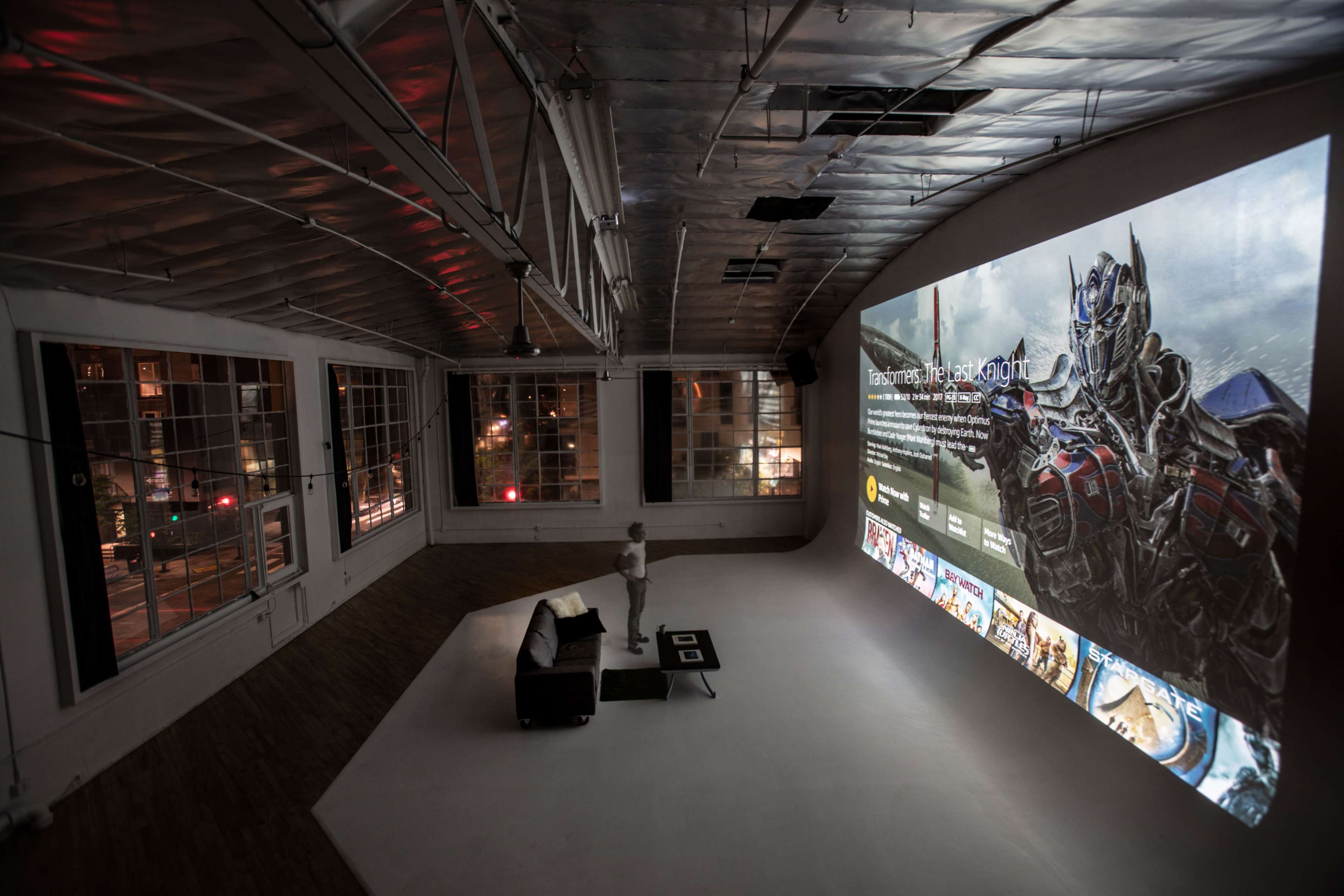 A person stands in a spacious, dimly lit room with a large projection of a digital screen displaying various media and an image of a robot character on one wall.