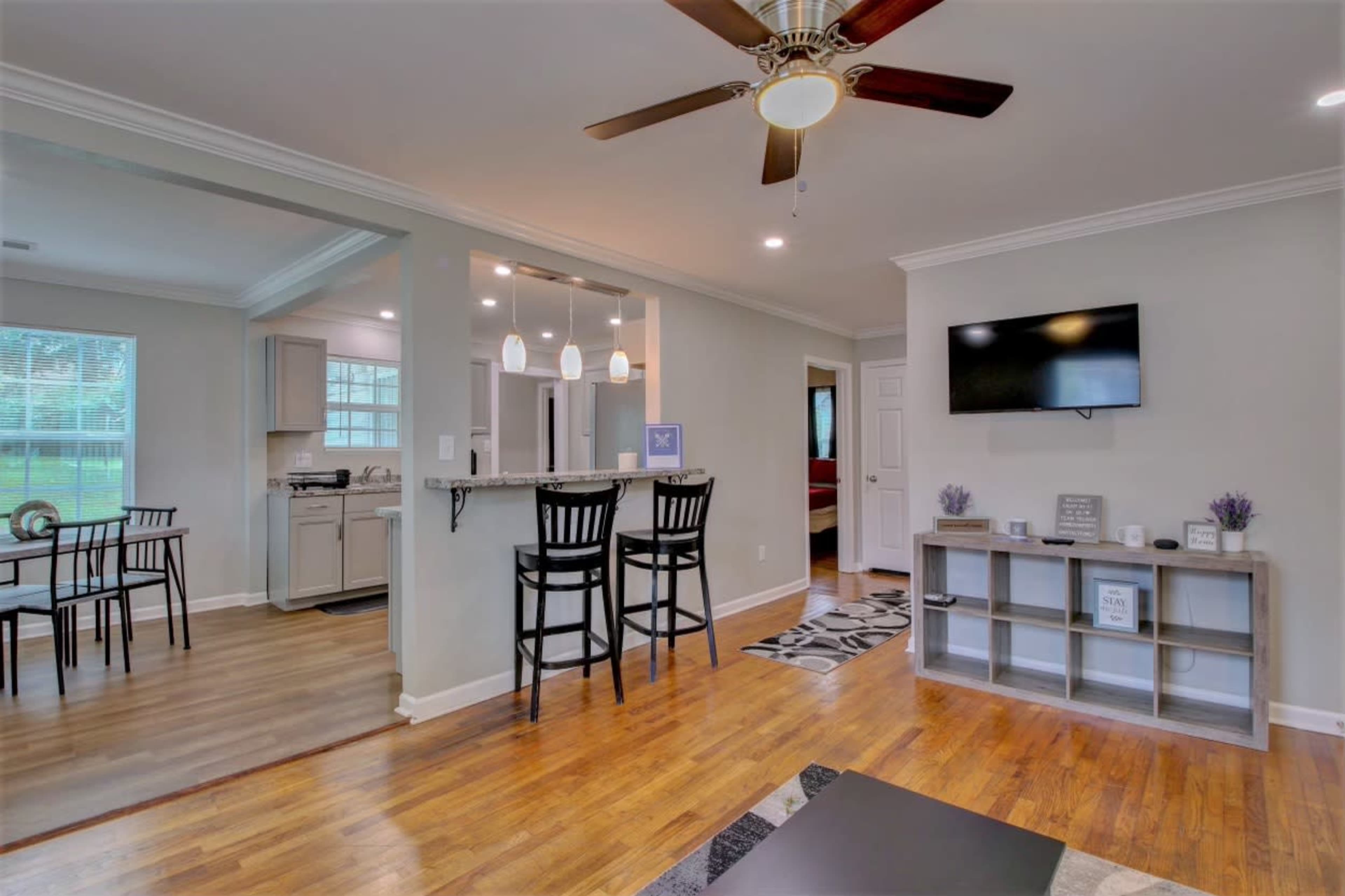 A room with hardwood flooring, a ceiling fan, a small bar area with two stools, and a television on the wall, with an adjacent open doorway leading to another room.