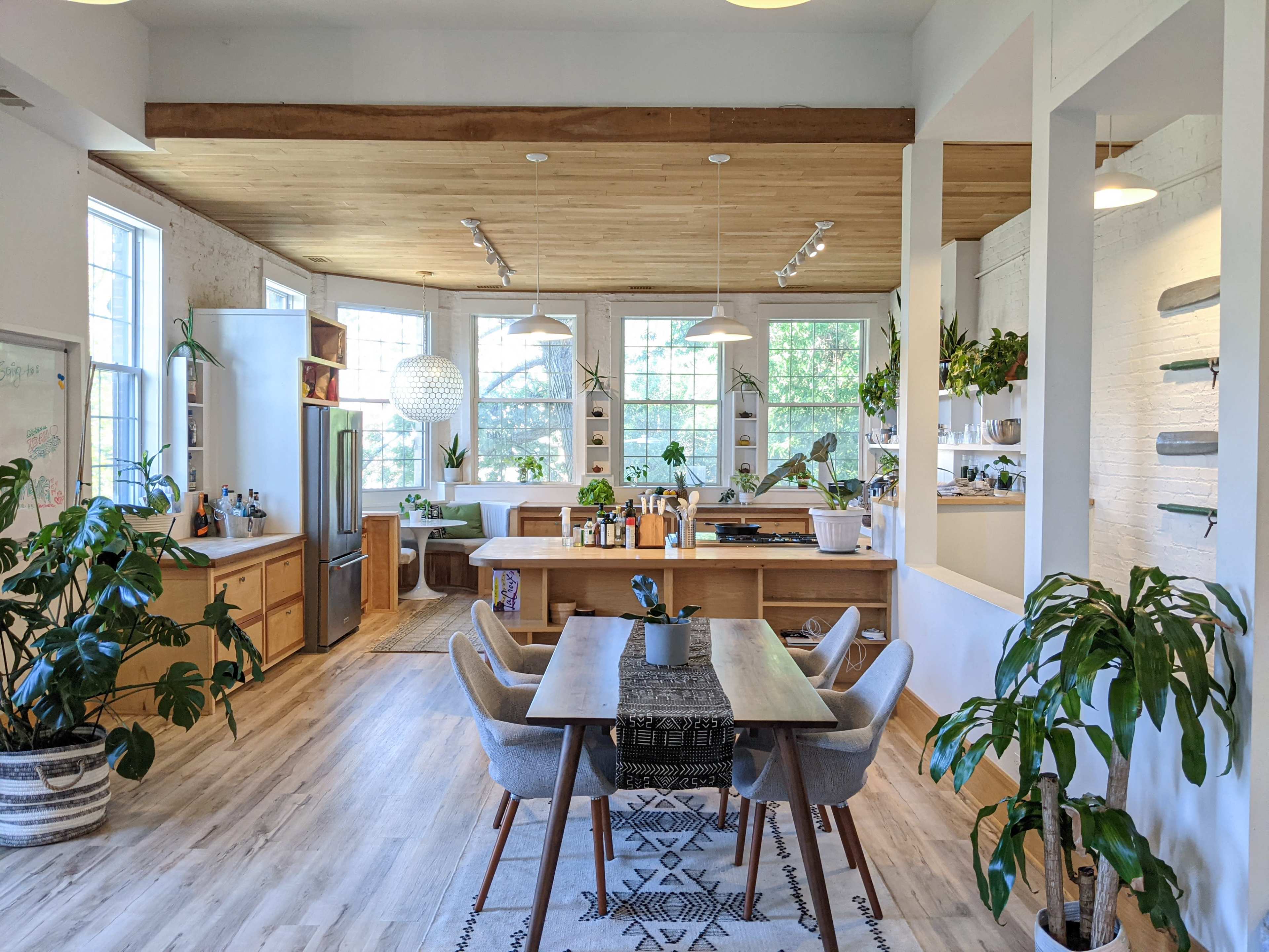The image shows a bright, modern kitchen and dining area featuring wooden cabinetry, a large dining table, and numerous houseplants.