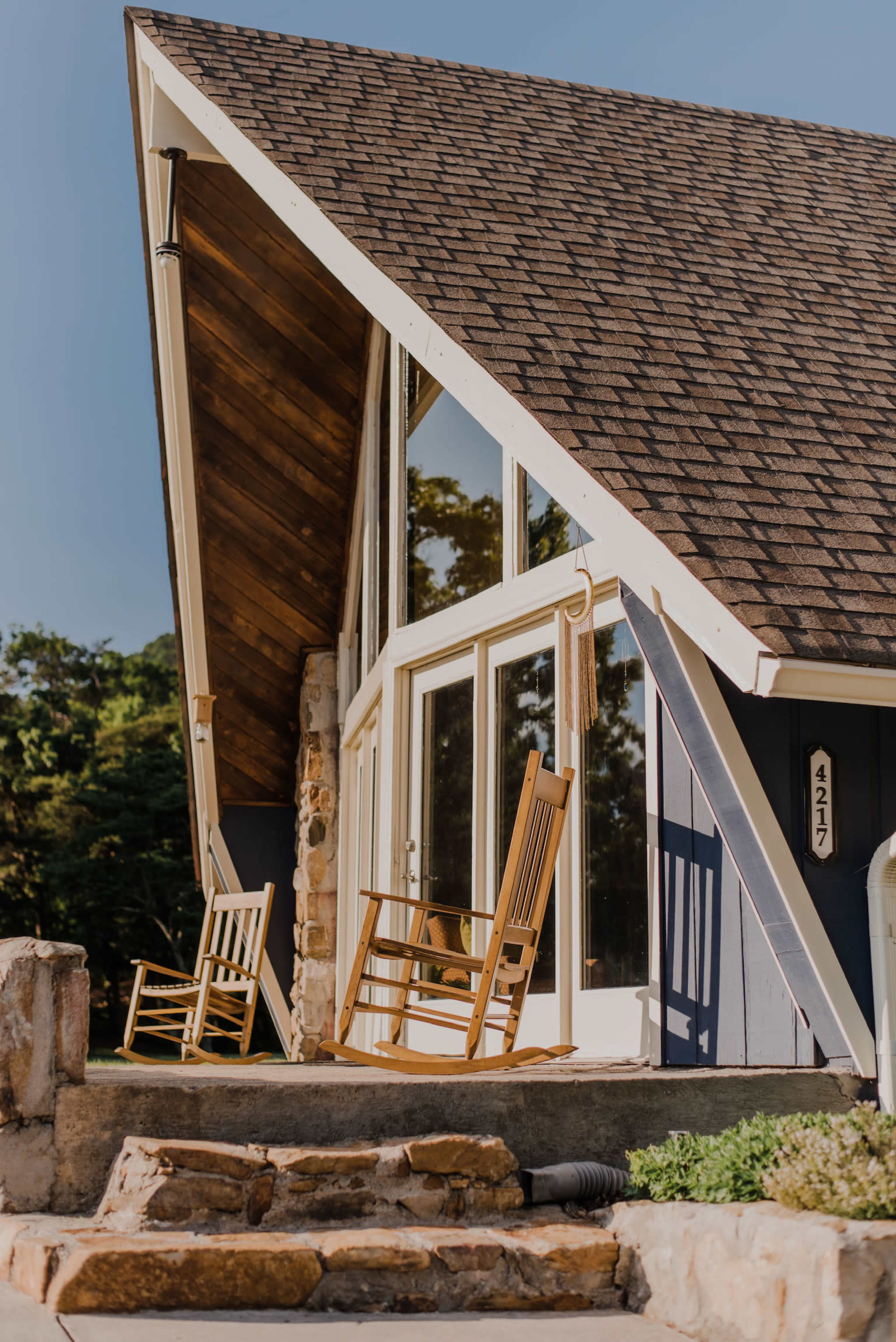 A modern A-frame house with large windows, two rocking chairs on a stone porch, and a stone pathway leading up to the entrance.