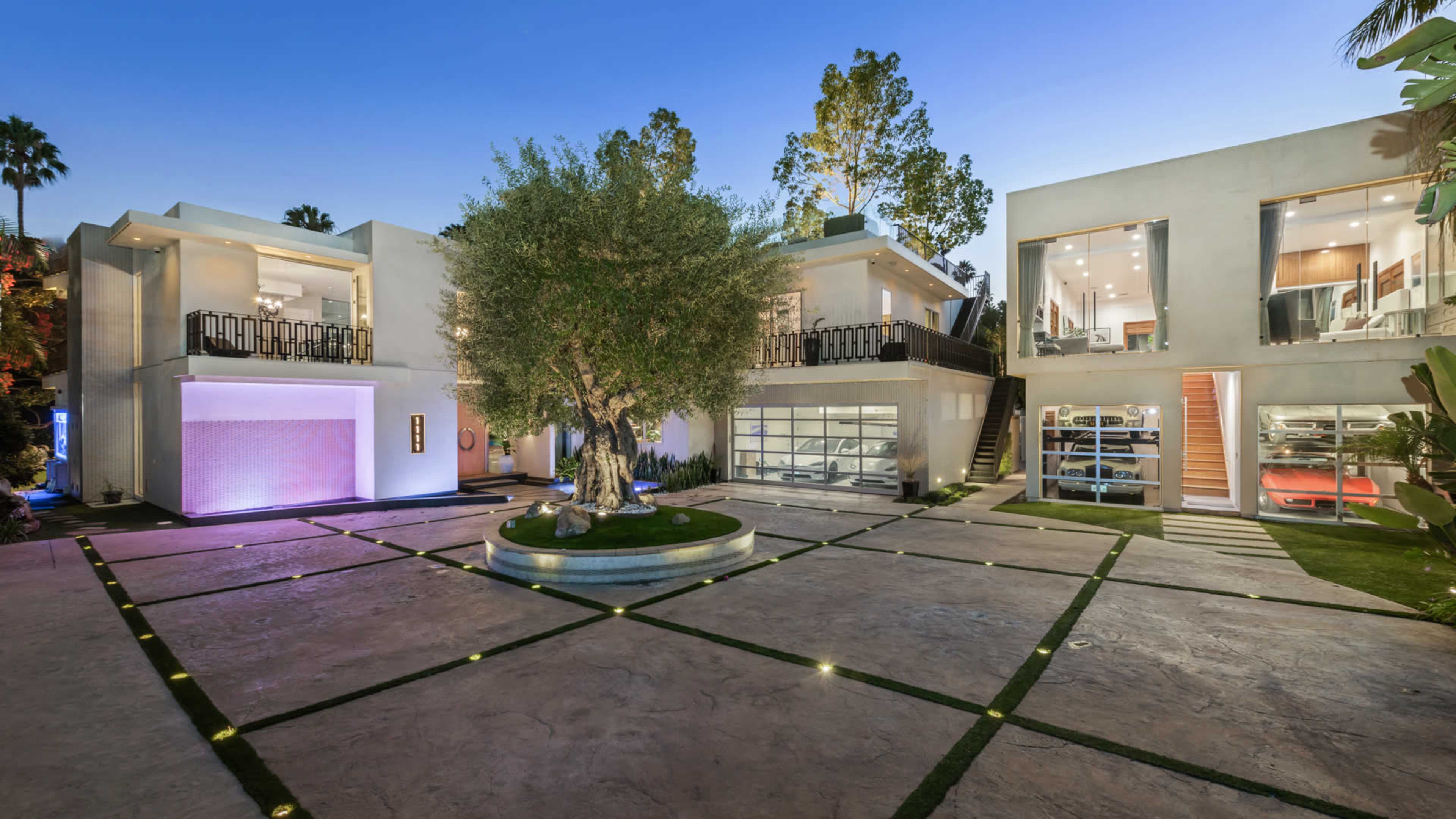 The image shows a modern, minimalist house surrounded by a landscaped courtyard with a large tree and a driveway leading to garage doors.