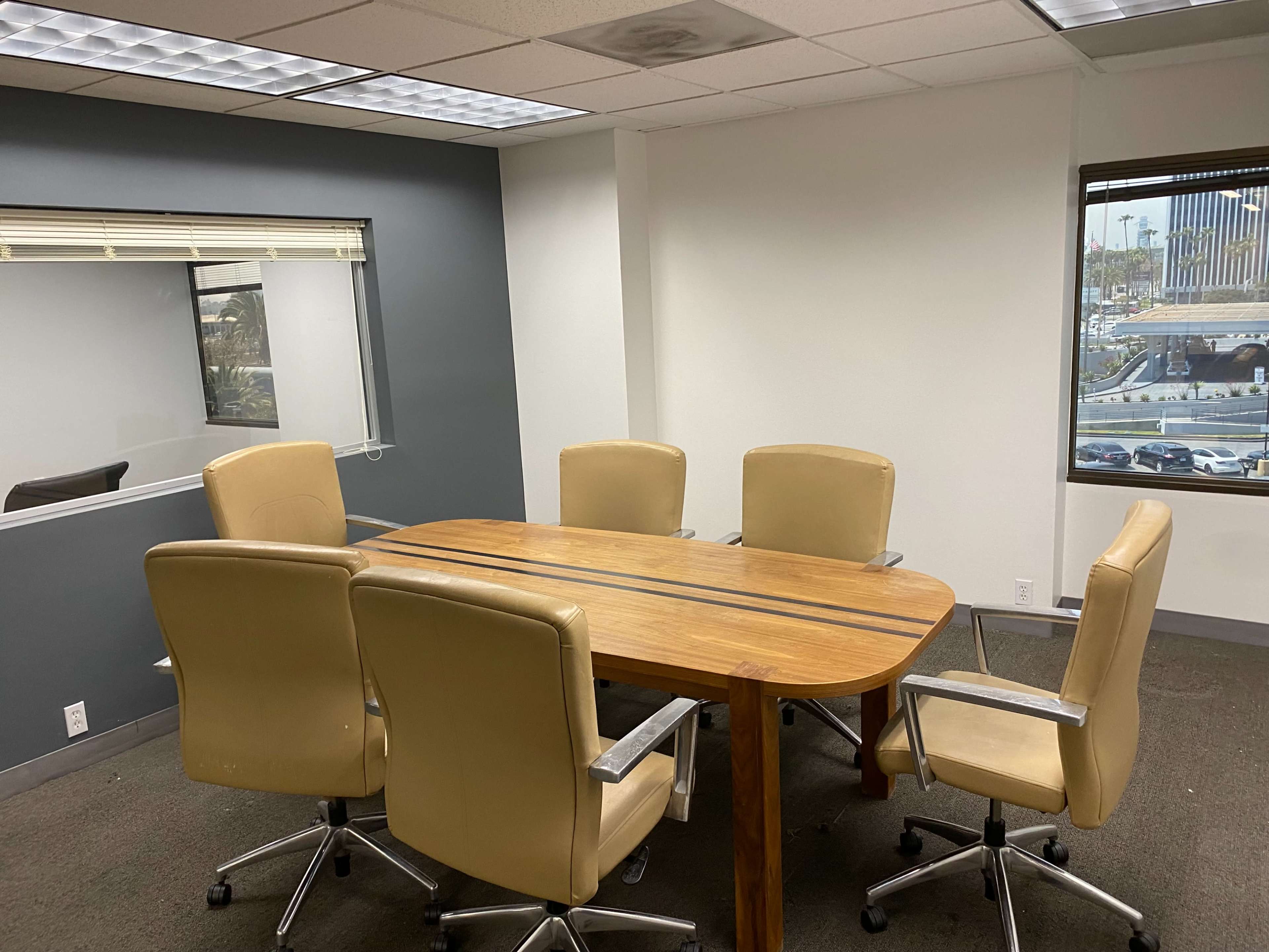 A conference room features a wooden table surrounded by five beige chairs and a large window with a view outside.