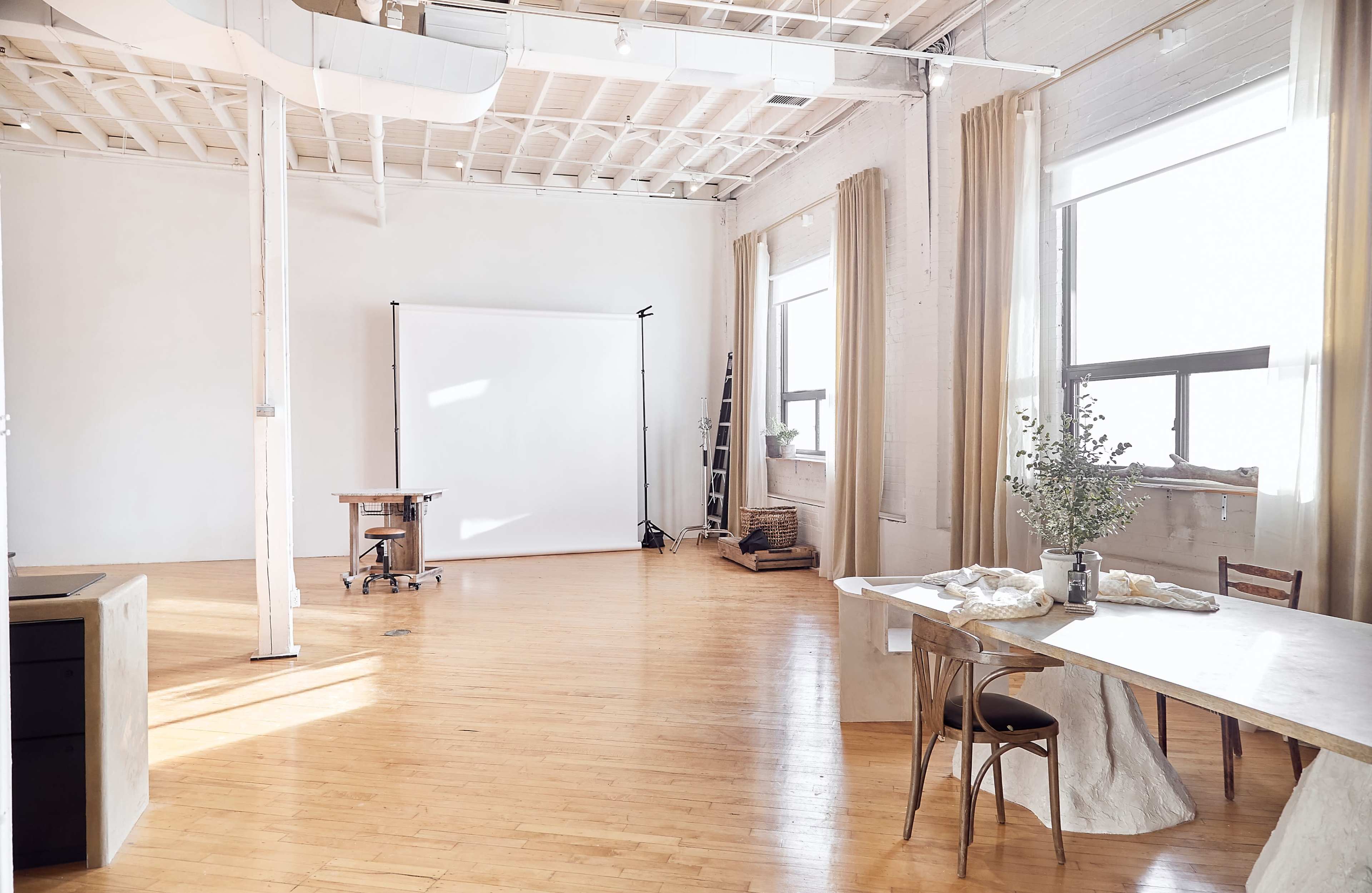 A bright, minimalist studio space featuring a large empty backdrop, wooden furniture, and large windows allowing natural light to fill the room.