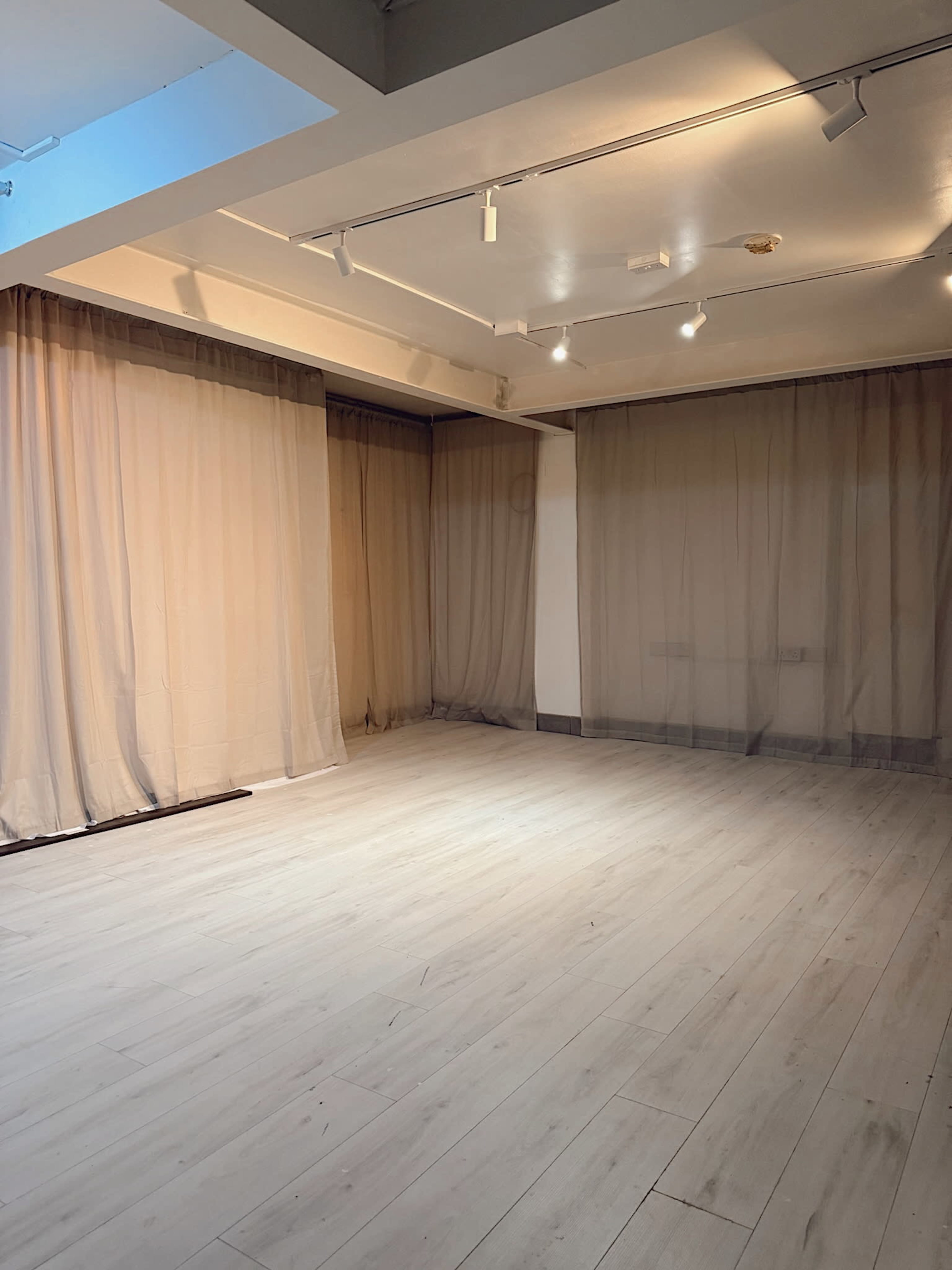 The image shows an empty room with light wood flooring, white walls, and sheer curtains hanging from the ceiling.