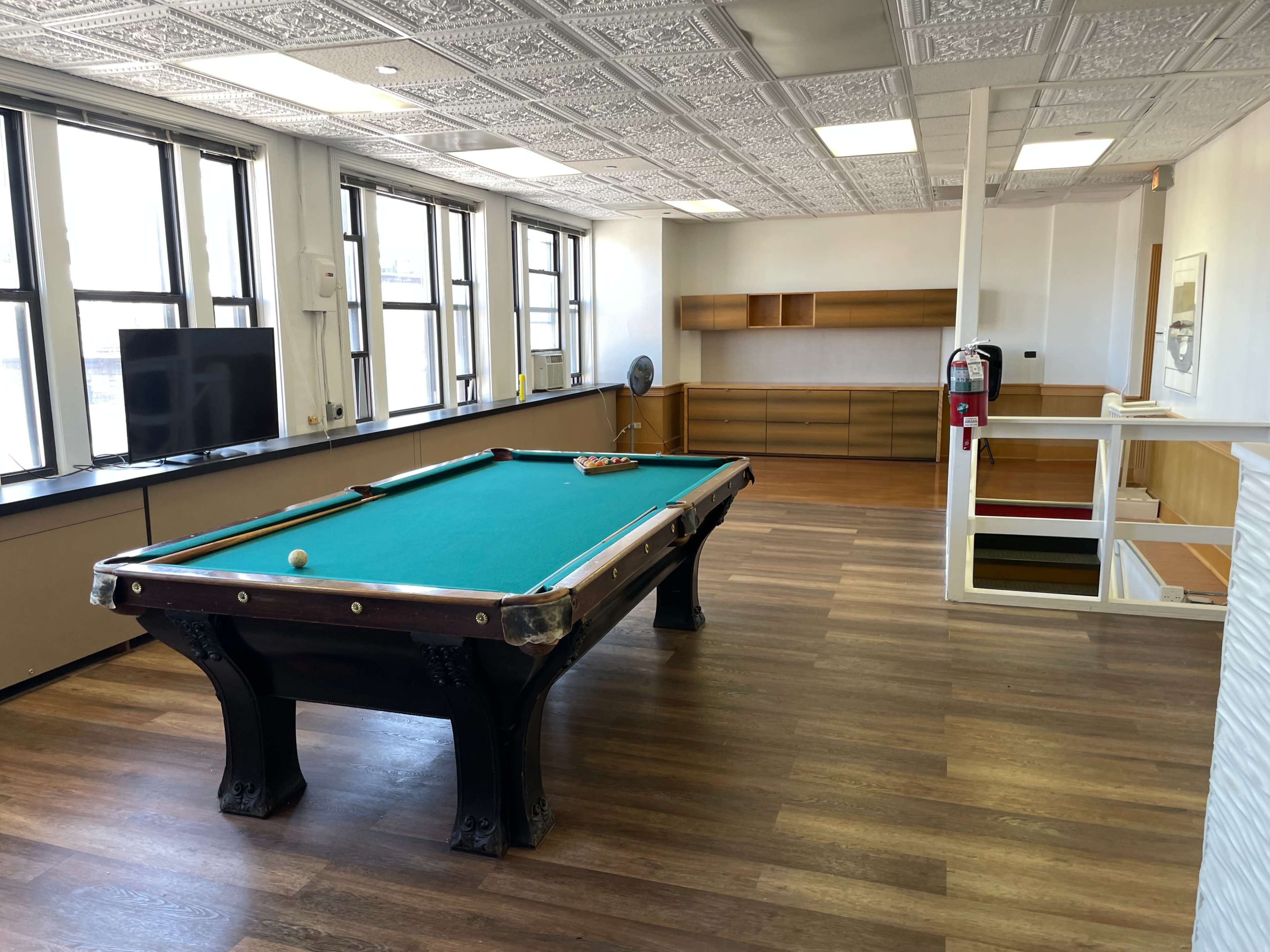 The image shows a spacious room with a pool table, large windows, and a wooden storage unit along one wall.