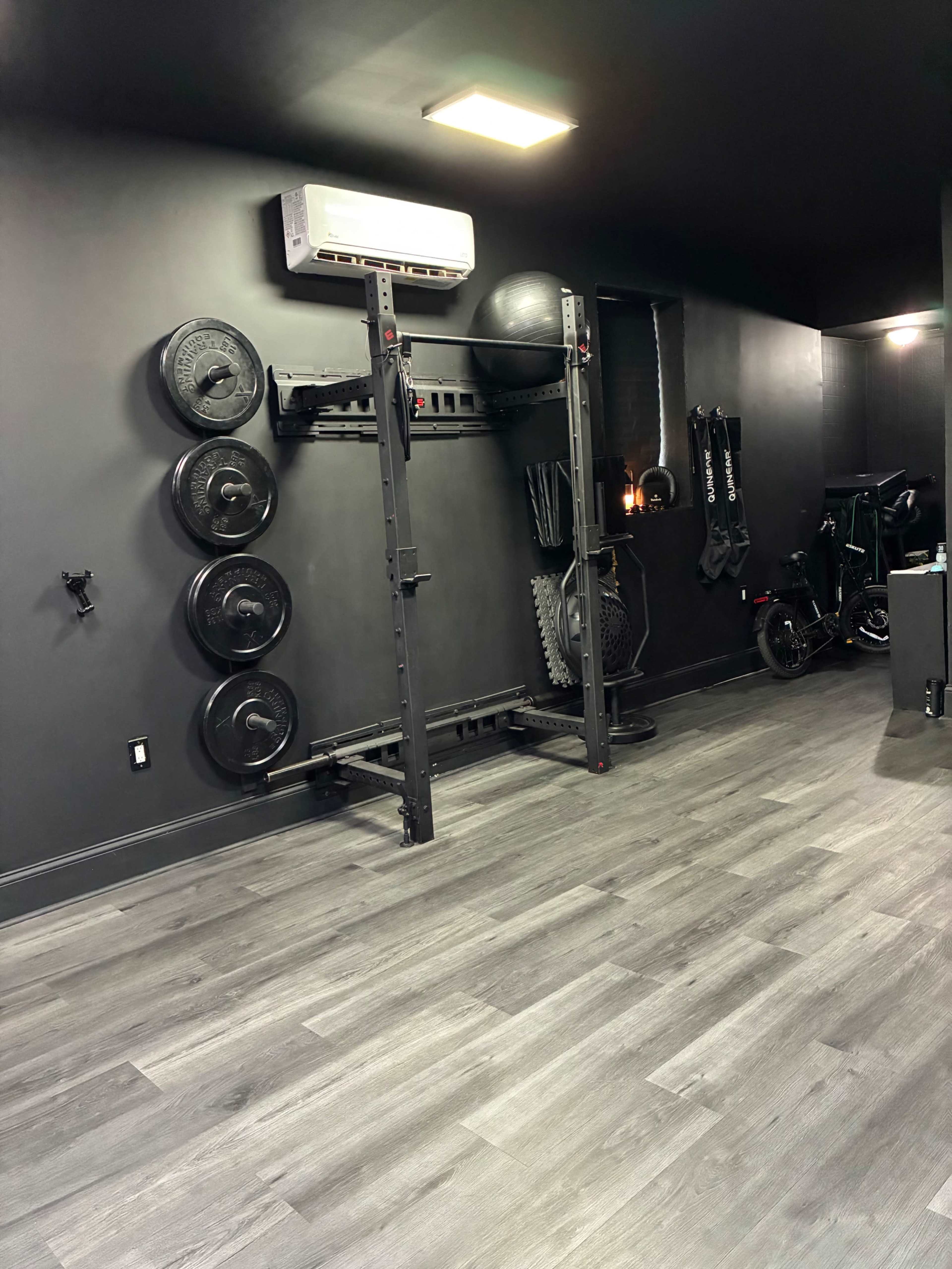 The image shows a dark-colored gym interior with various weight plates, a rack, and fitness equipment arranged along one wall.