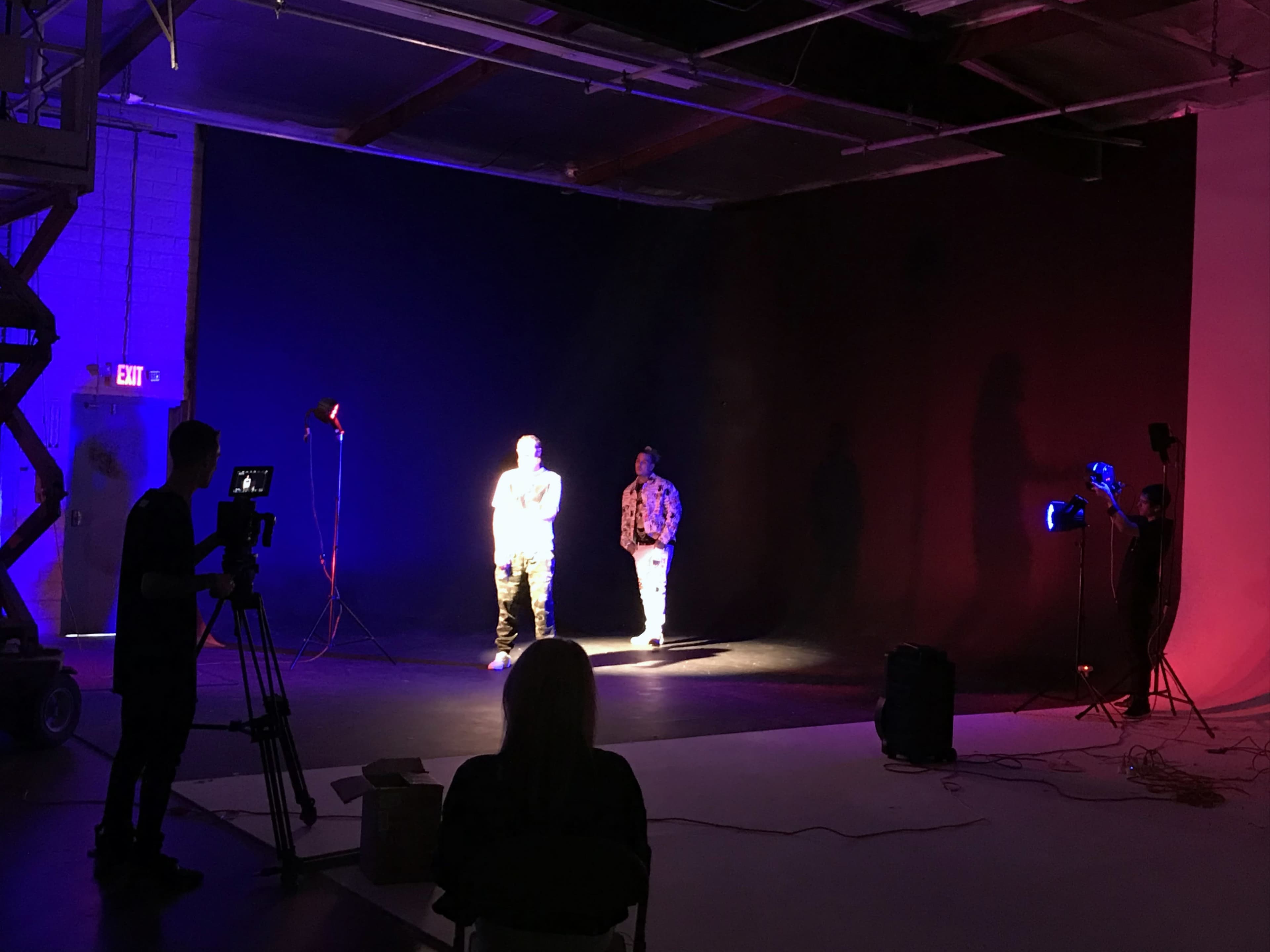 Two individuals are illuminated in a dimly lit studio, surrounded by various stage lights and camera equipment.