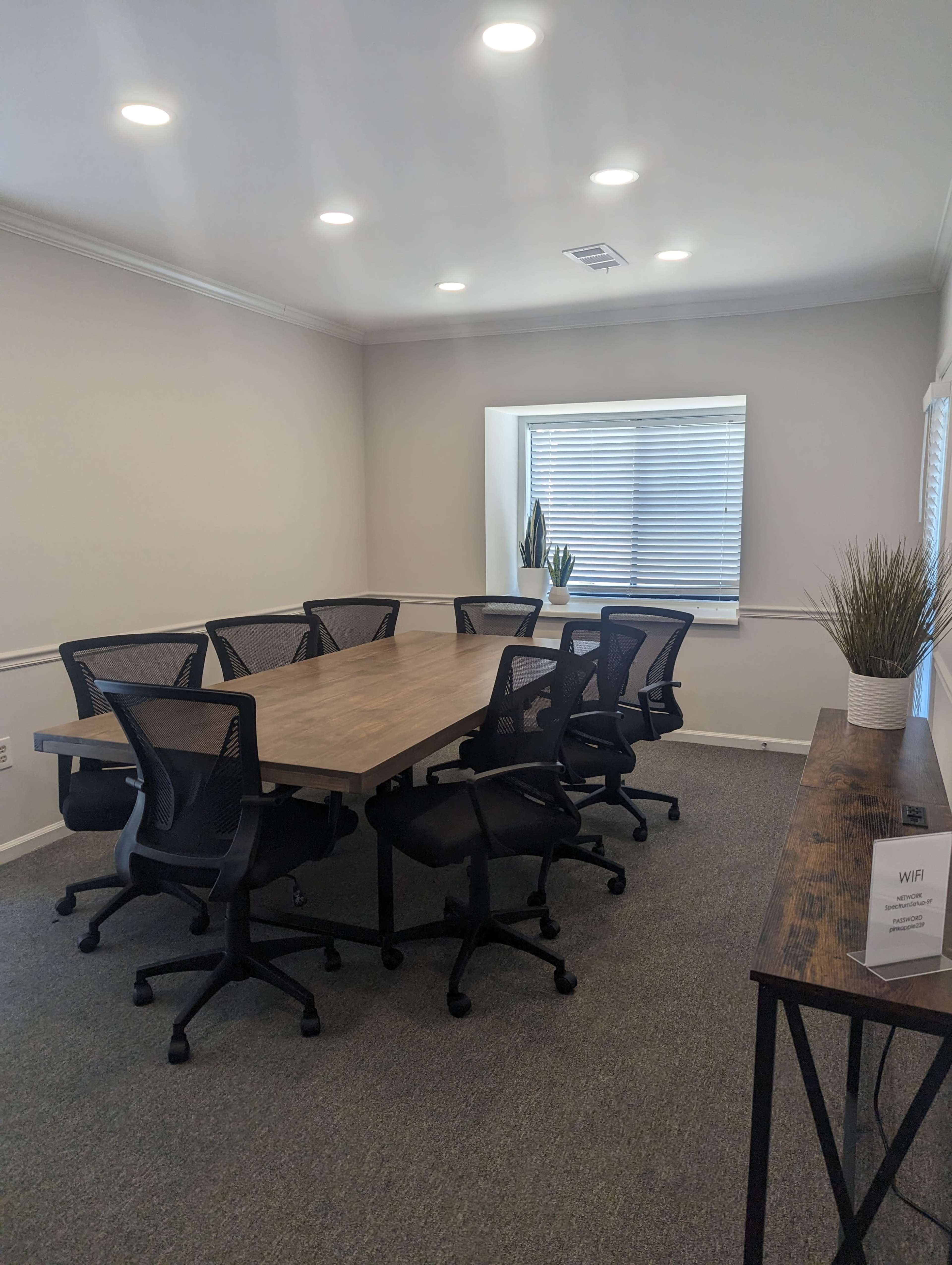 A conference room features a long wooden table surrounded by eight black office chairs, with a window and a plant in the background.
