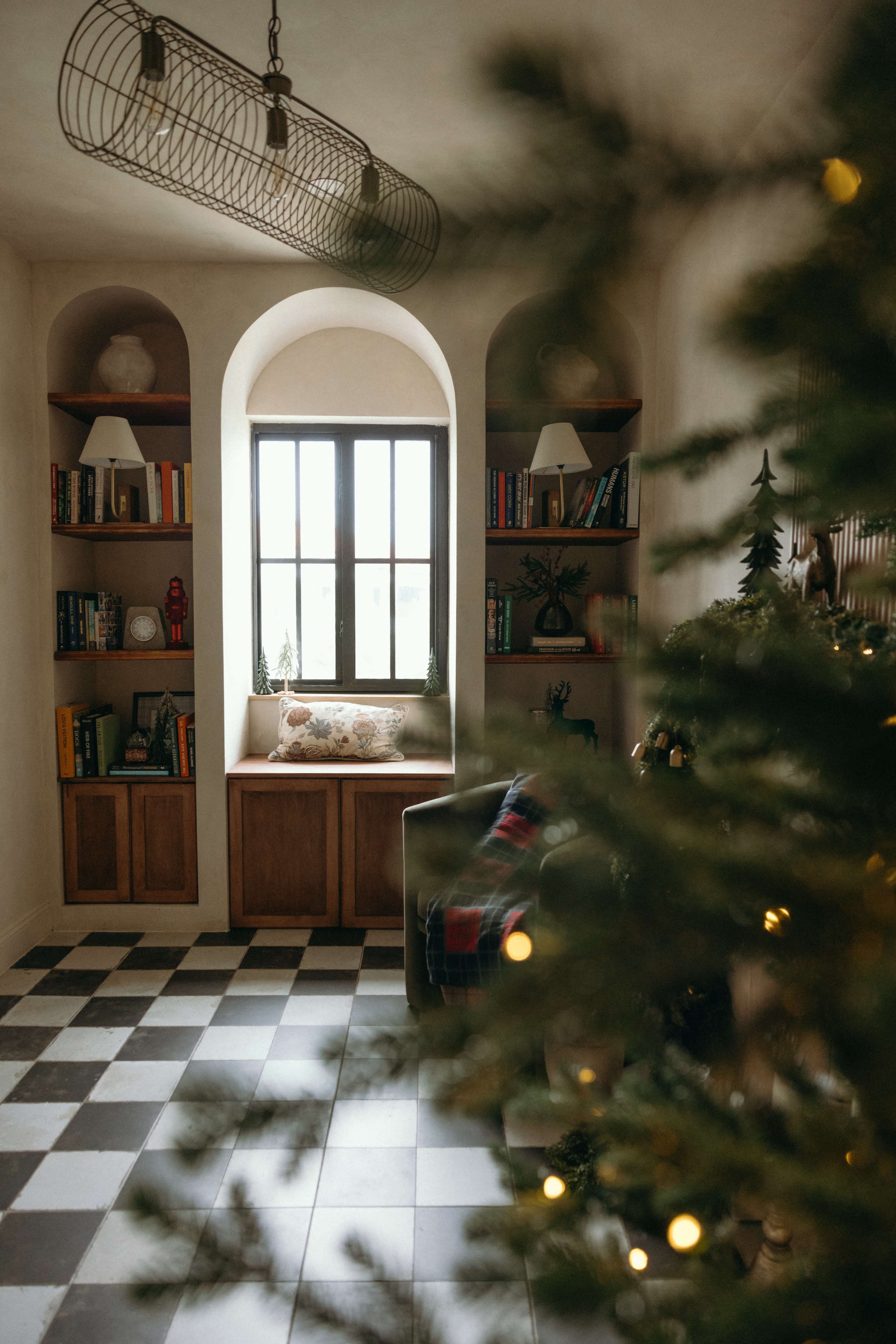 The image features a cozy interior room with a large window, bookshelves filled with books, and a decorated Christmas tree in the foreground.