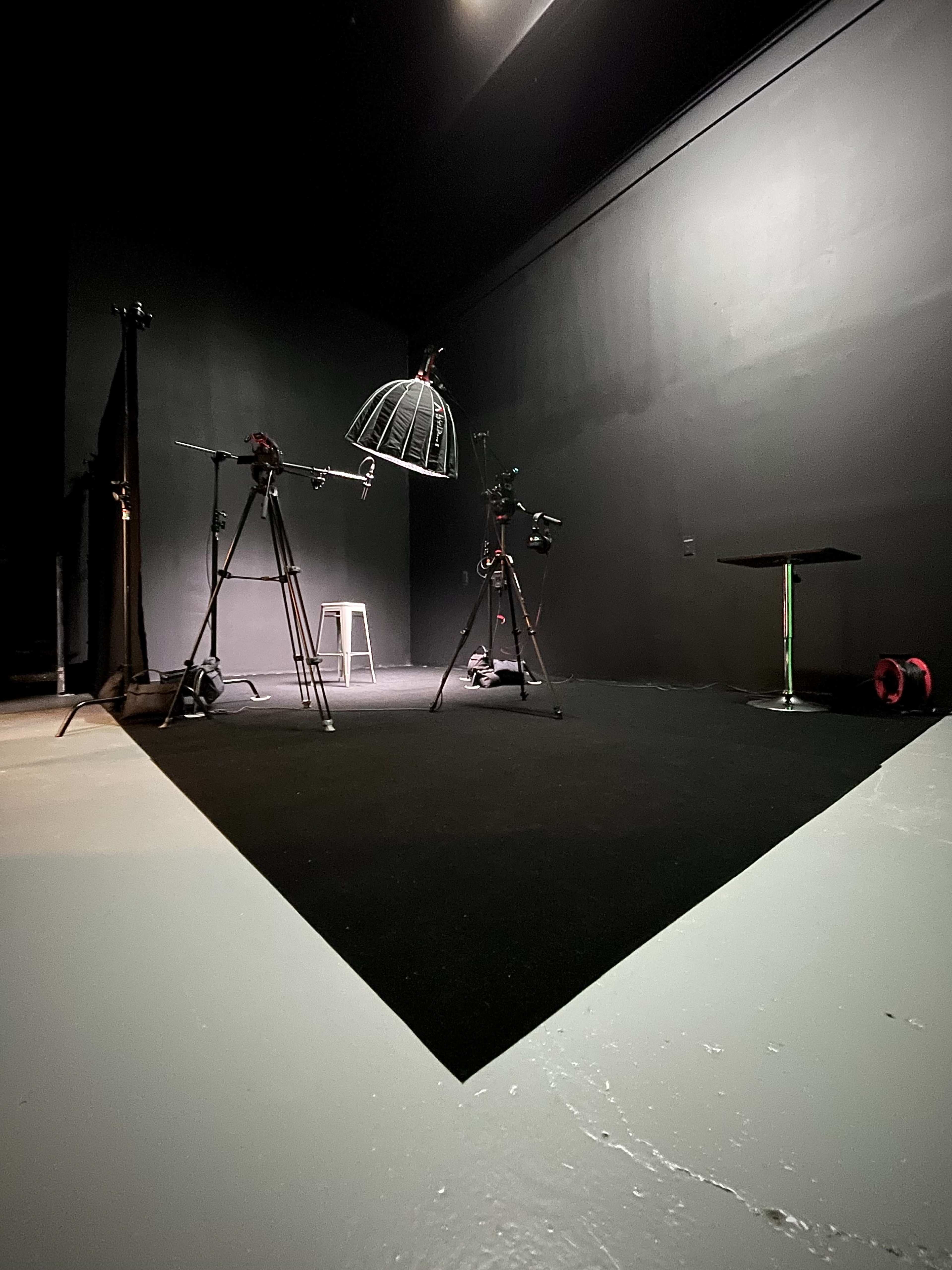 The image shows a dark studio space with two camera tripods, a light fixture, and a small table on a black floor.