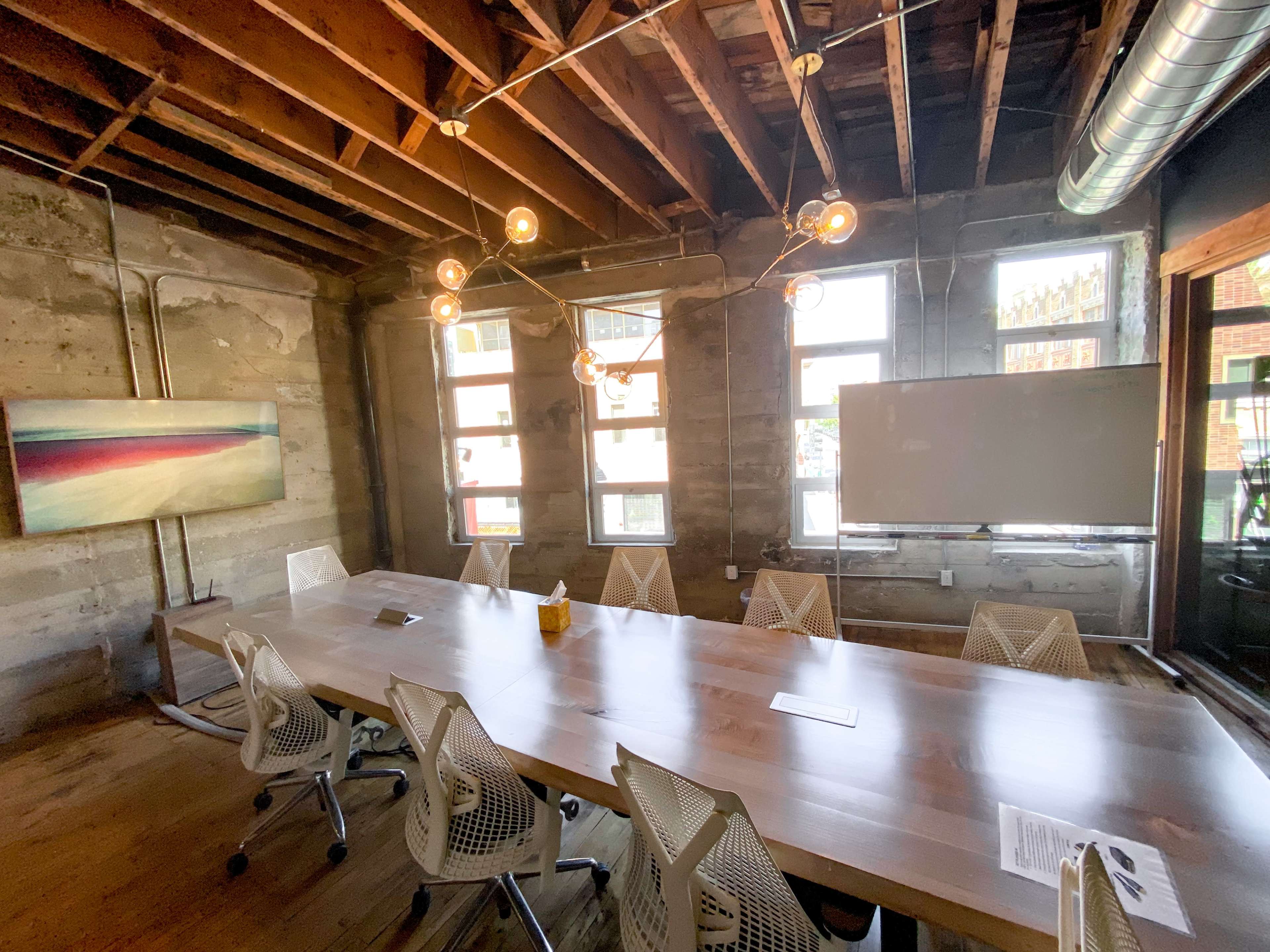 A modern conference room features a long wooden table surrounded by ergonomic chairs, with exposed beams and large windows letting in natural light.