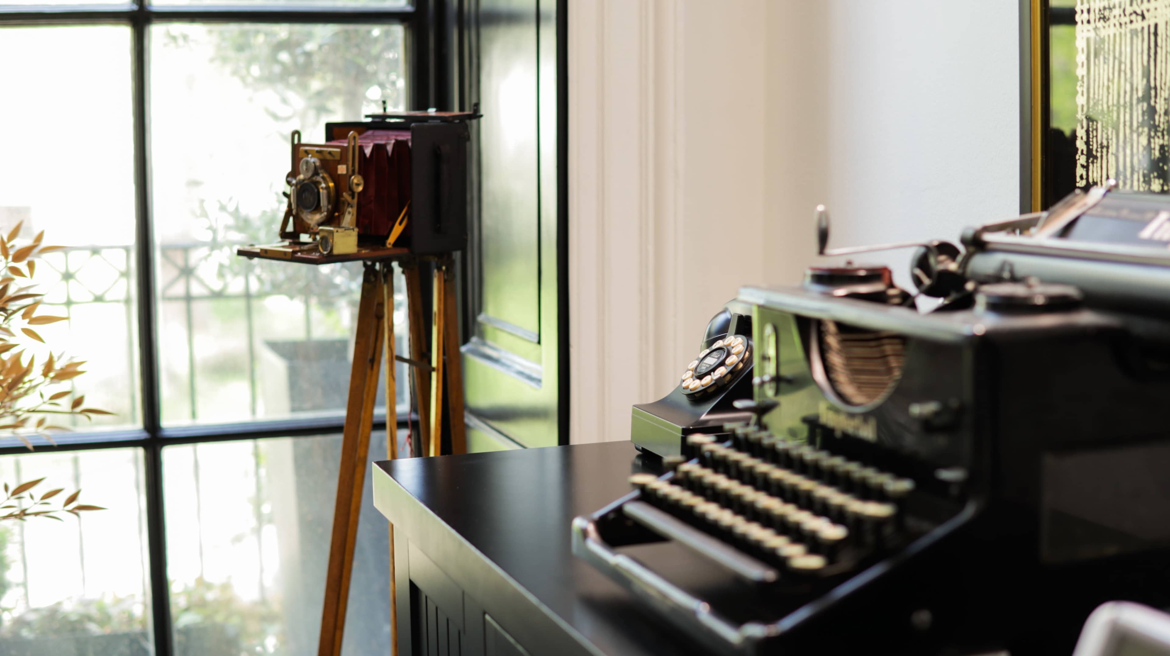 The image shows a vintage typewriter and an old-fashioned camera on a table next to a window.
