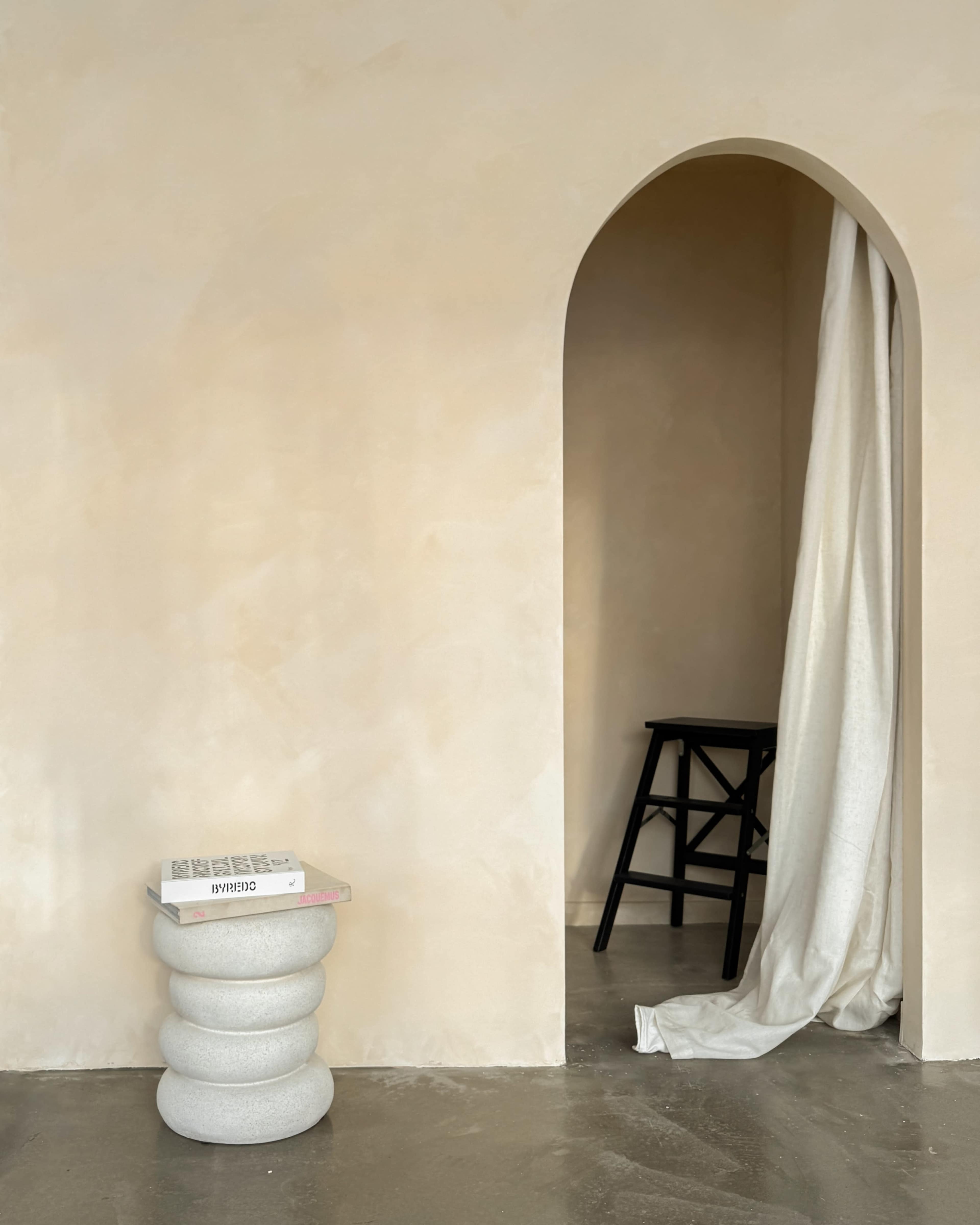 A minimalist interior features a stone-colored wall, a curved archway, a black wooden stool, and a soft white fabric draped nearby, with a sculptural pedestal holding a book.