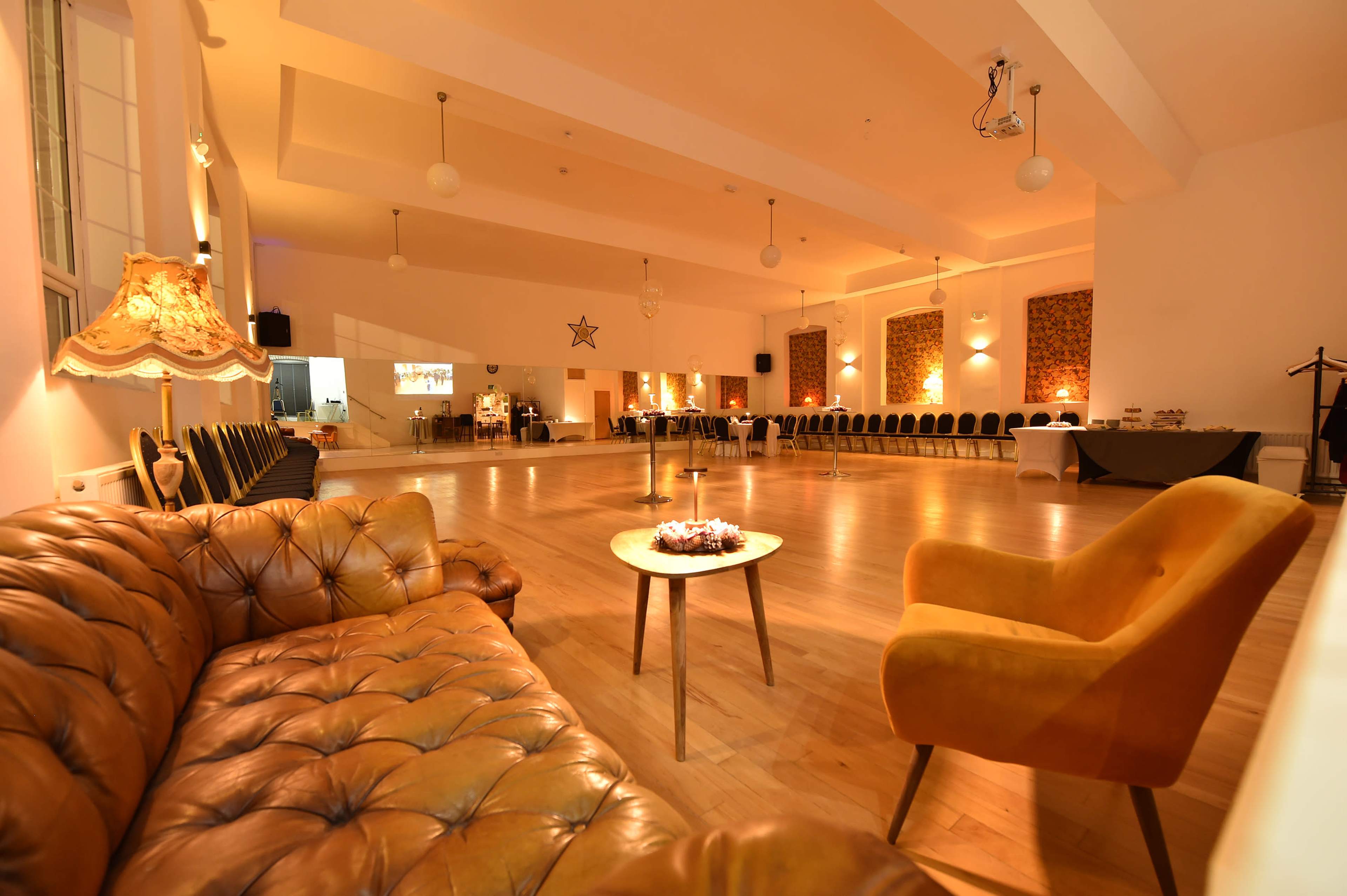 The image shows a spacious event room with a polished wooden floor, a few round tables, and a cozy seating area featuring a brown leather couch and a yellow armchair.