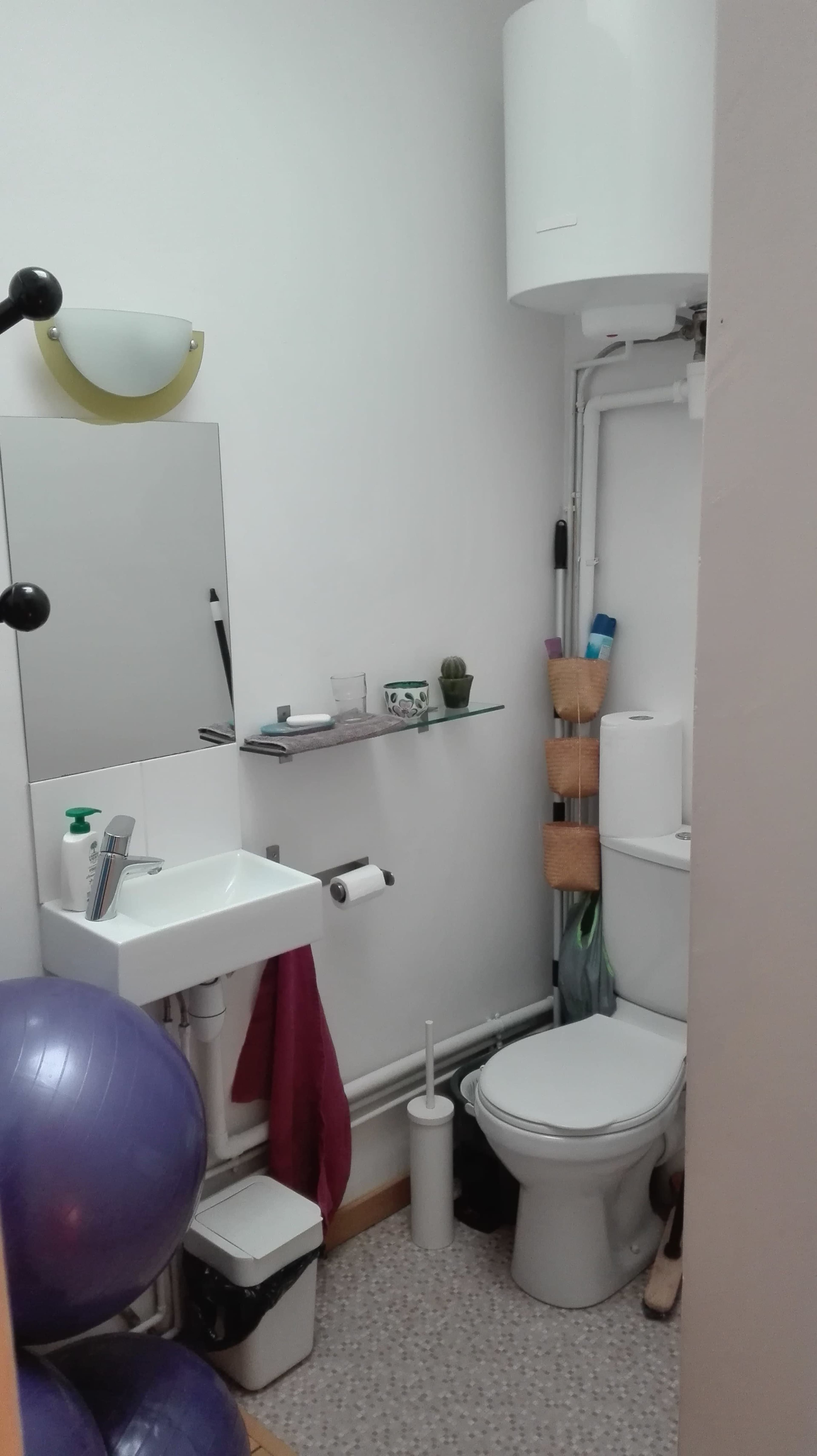 A small bathroom with a sink, toilet, mirror, shelves, and exercise ball in the corner.