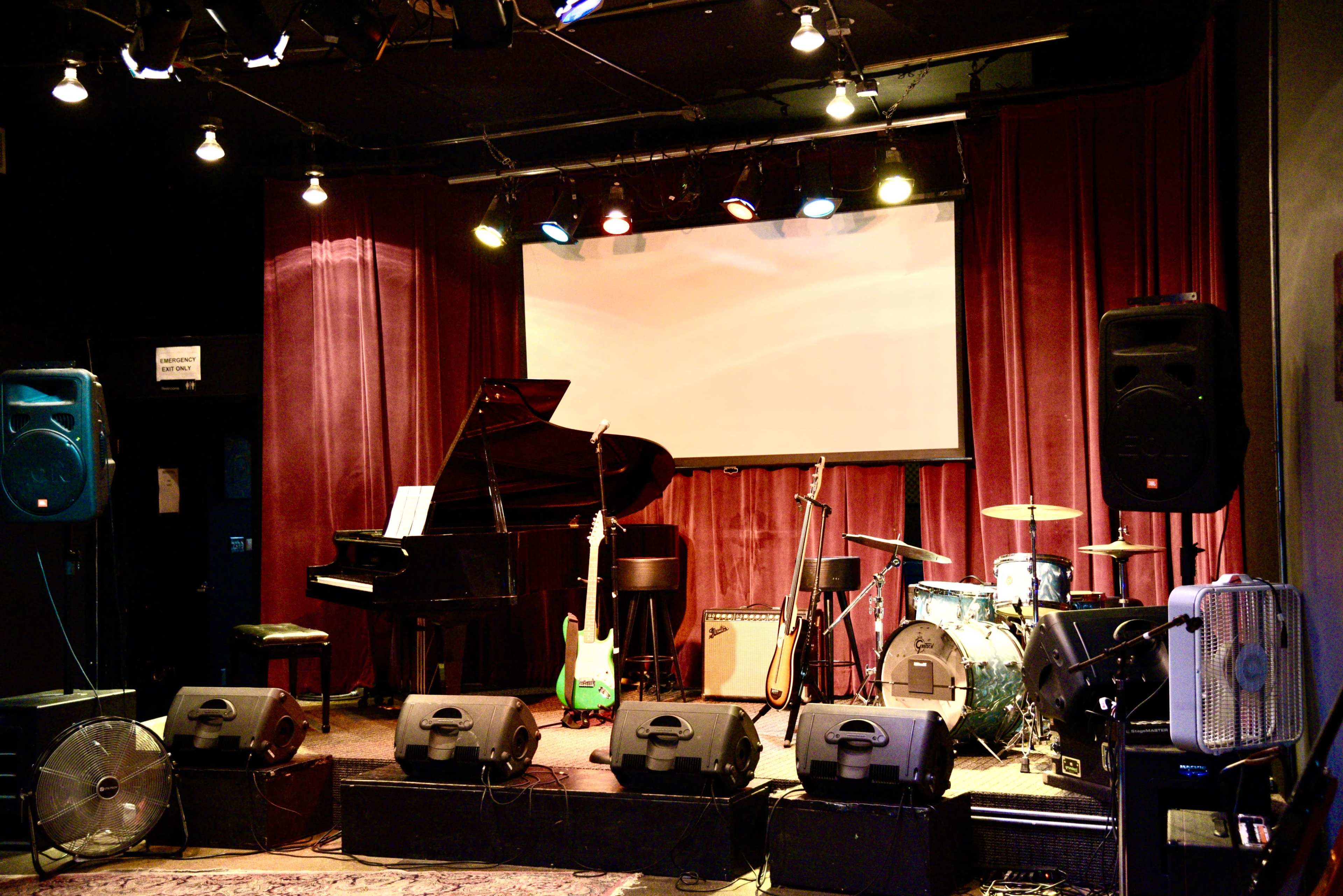 The image shows a stage set up for a performance, featuring a grand piano, microphones, guitars, a drum set, and various amplifiers, with red curtains in the background.
