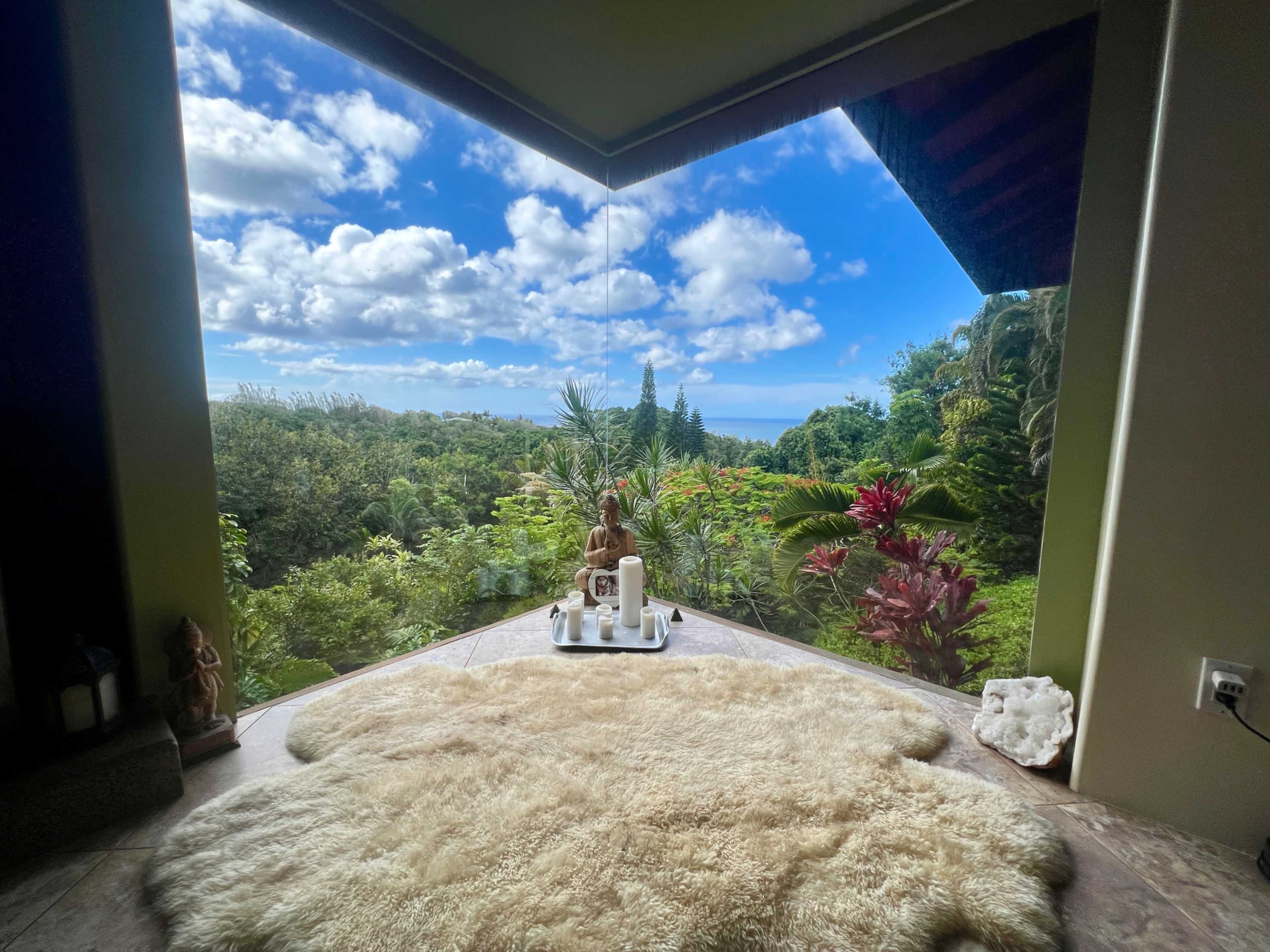 A serene interior space features a plush rug and a small altar with a statue and candles, framed by large windows overlooking a lush, green landscape under a partly cloudy sky.