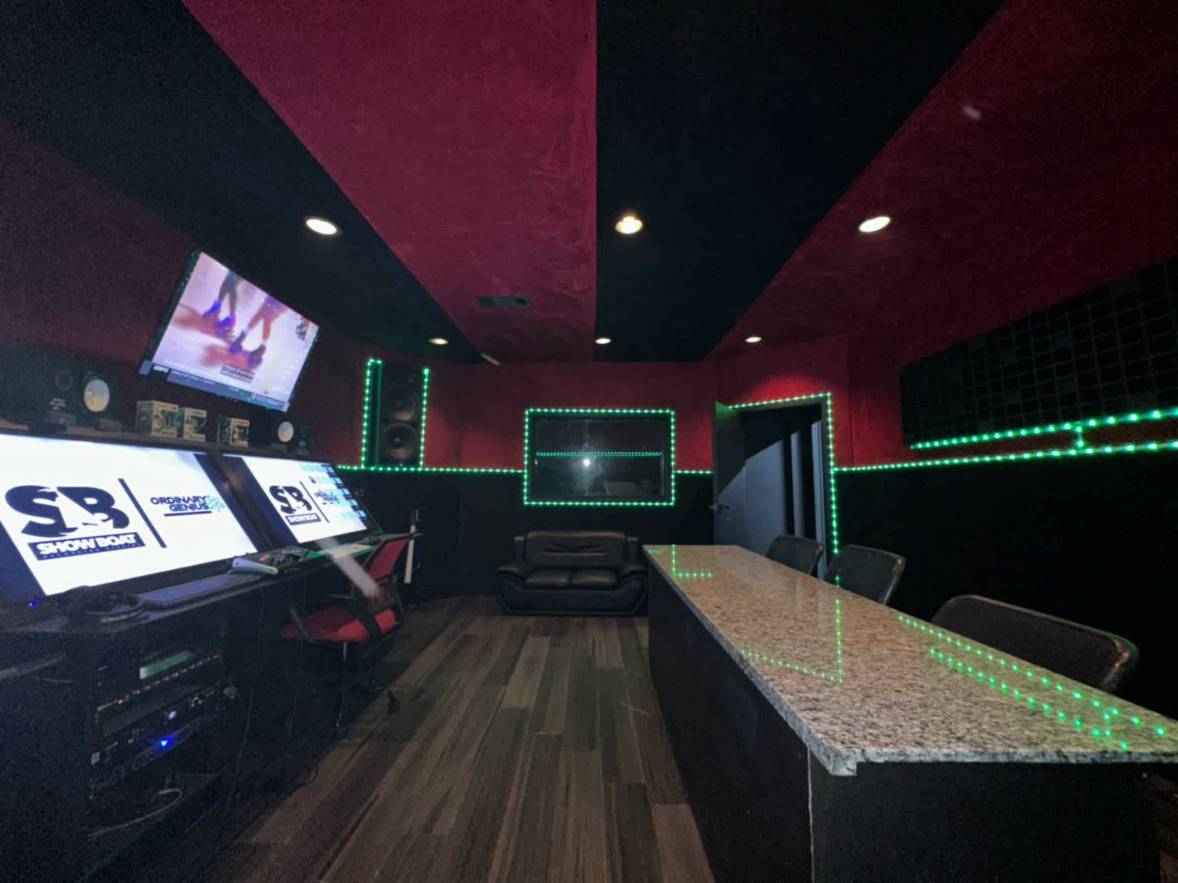 The image shows a music studio with dark walls, a granite countertop, several computer monitors displaying logos, and green LED lights outlining the space.