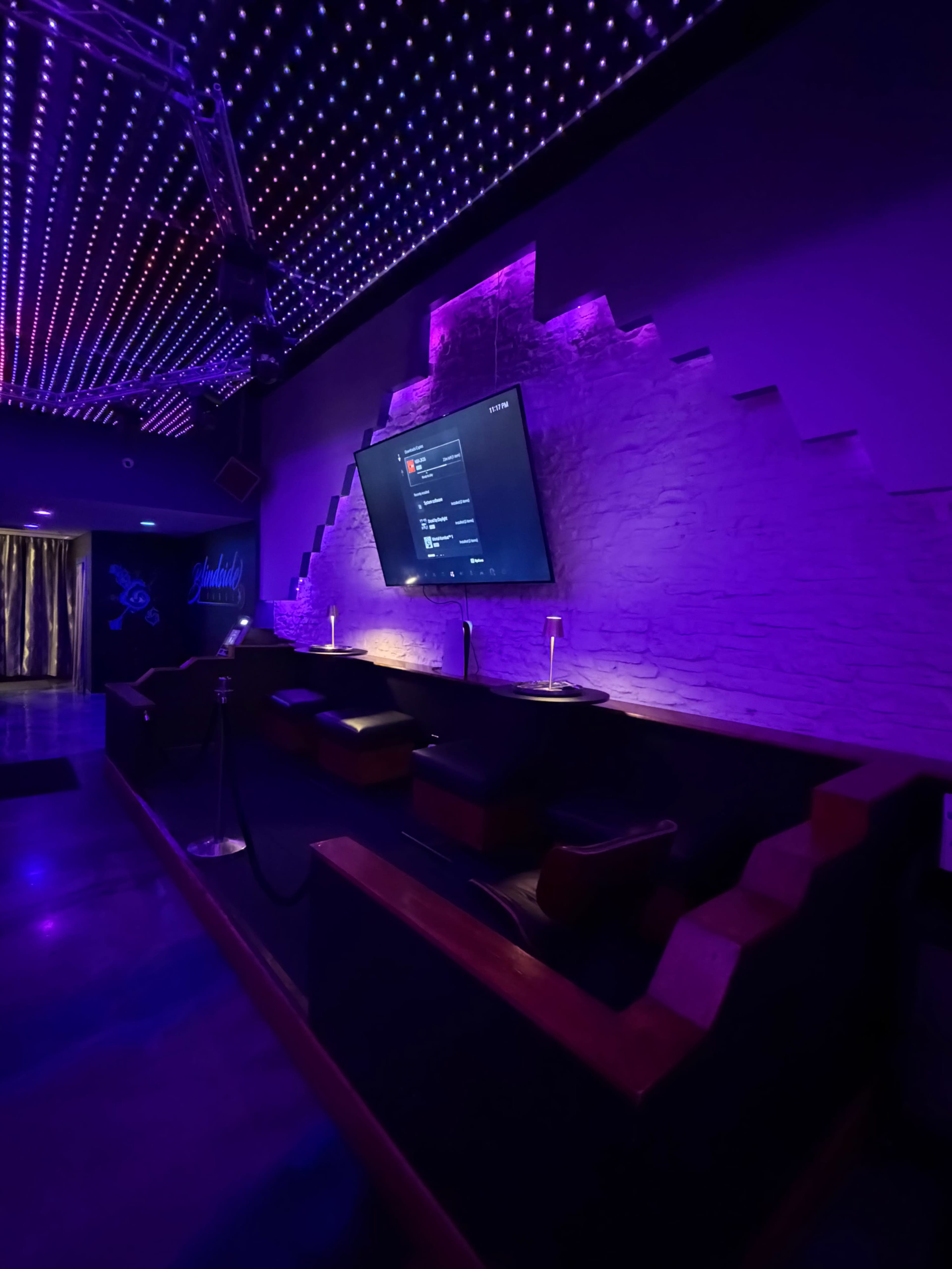 The image shows a modern lounge area with purple LED lighting, a large flat-screen TV mounted on a textured wall, and several seating arrangements.