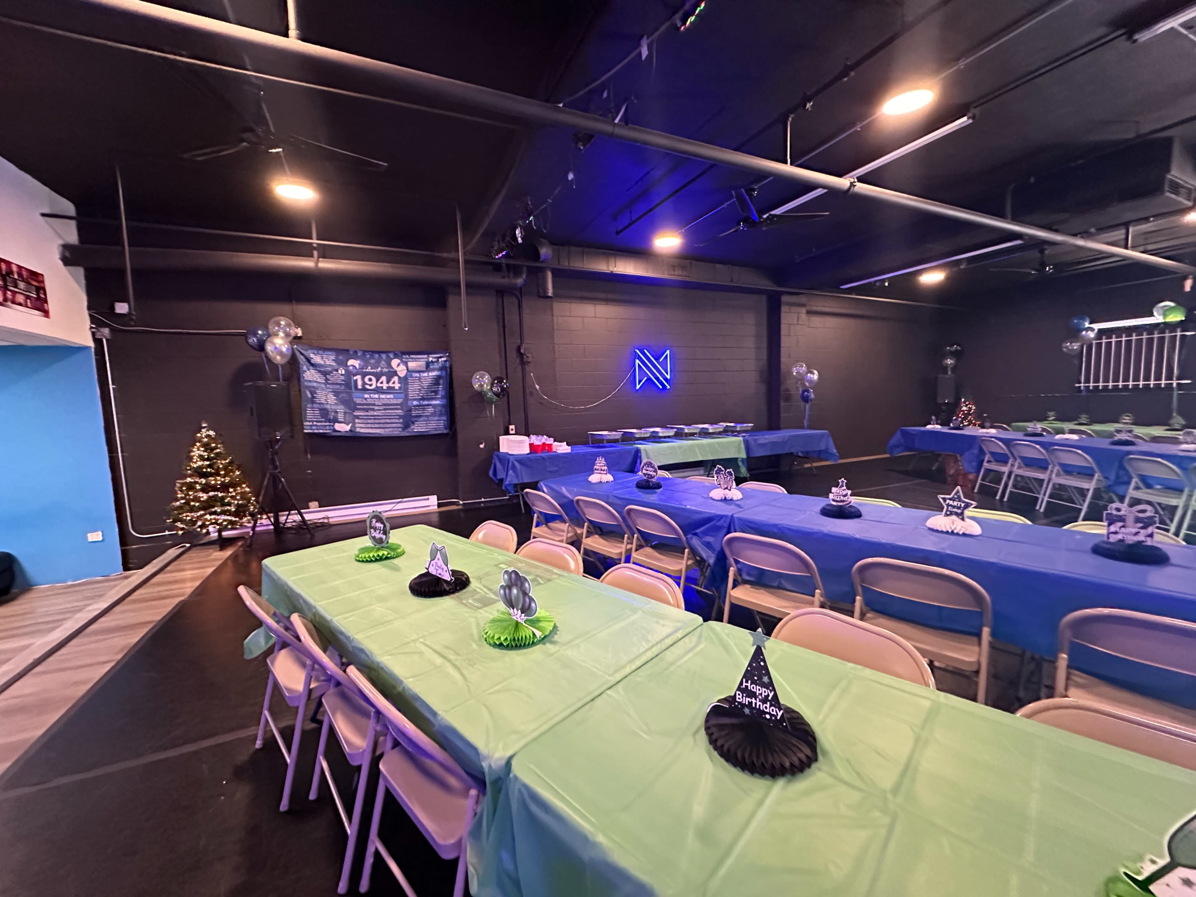 A spacious event room is set up with tables covered in blue and green tablecloths, decorated with hats and centerpieces, and a tree is visible in one corner.