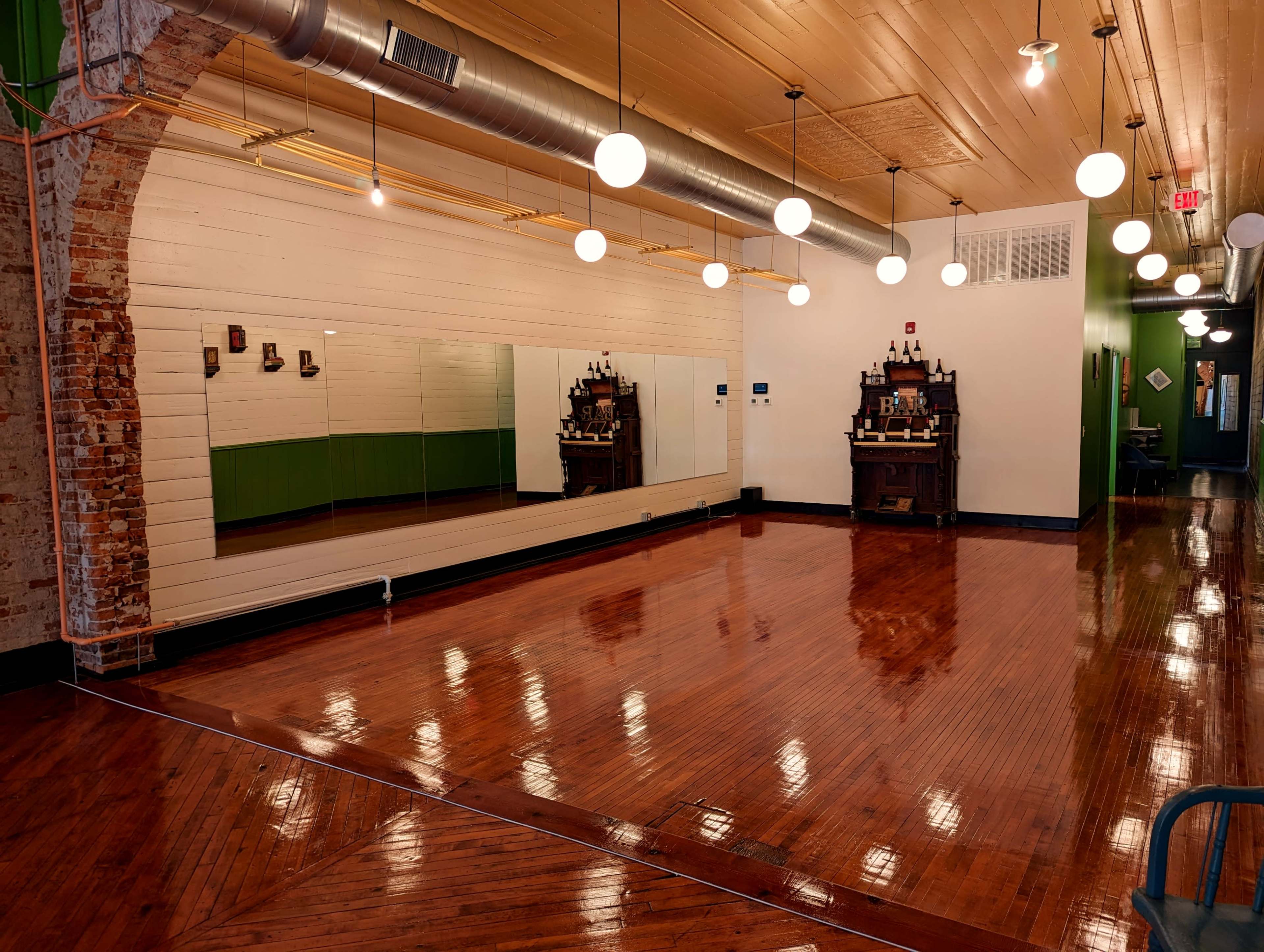A spacious room with polished wooden floors, large mirrors on one wall, and a decorative shelving unit against another wall, illuminated by overhead pendant lights.