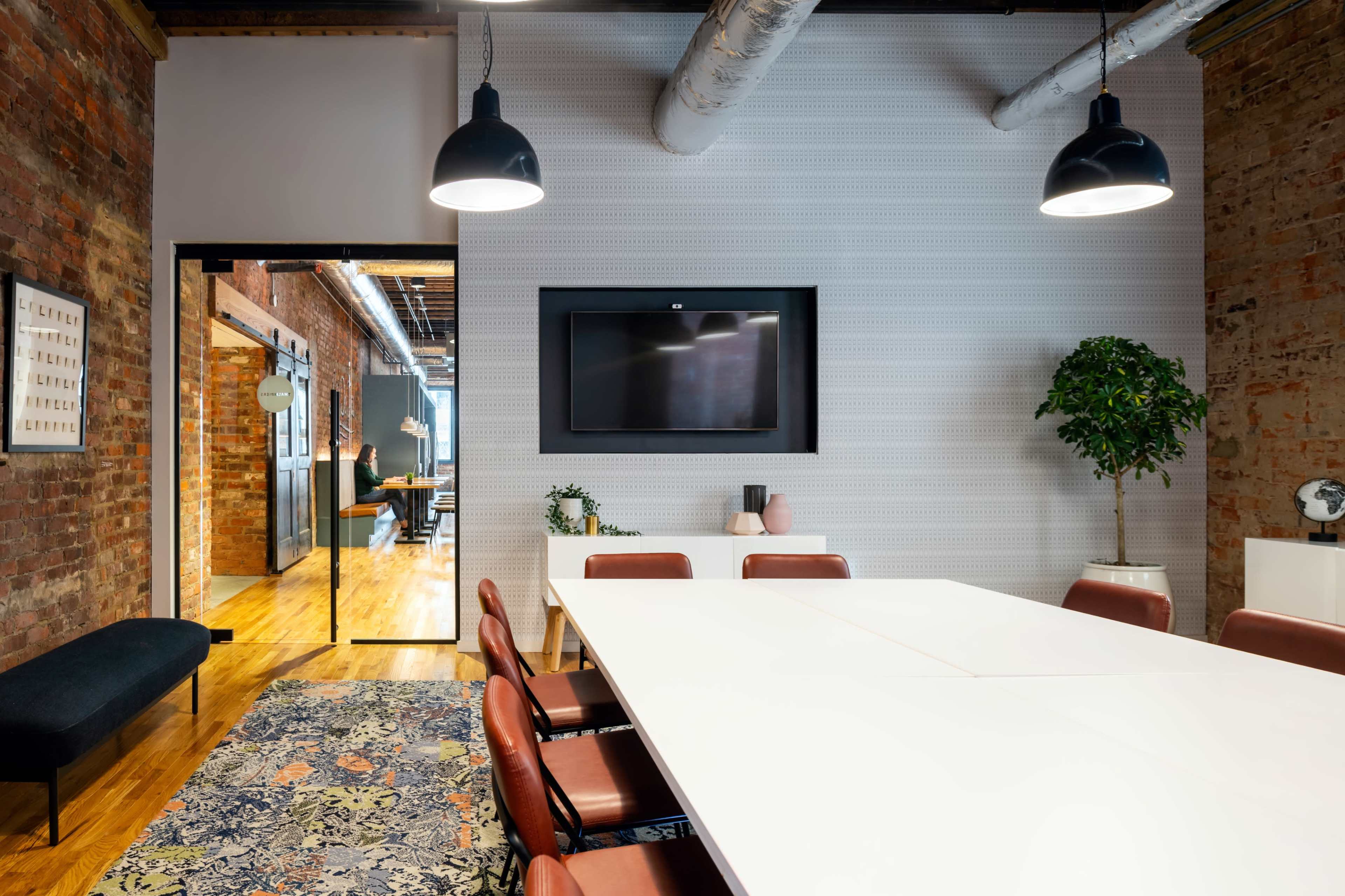 A modern conference room features a long white table surrounded by brown chairs, with exposed brick walls, a television mounted on the wall, and a decorative plant in the corner.