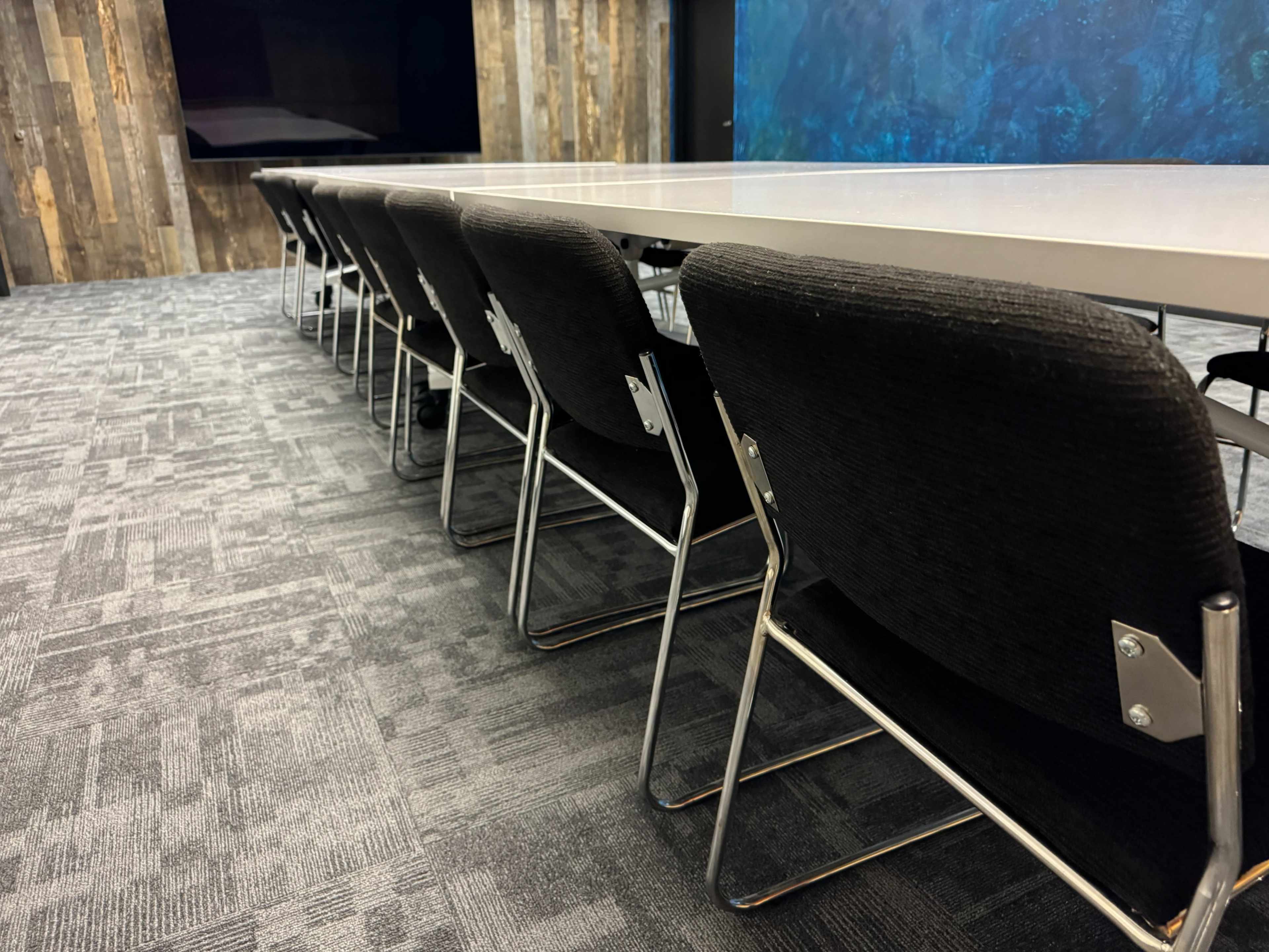 A long, empty conference table is surrounded by black chairs in a modern meeting room.