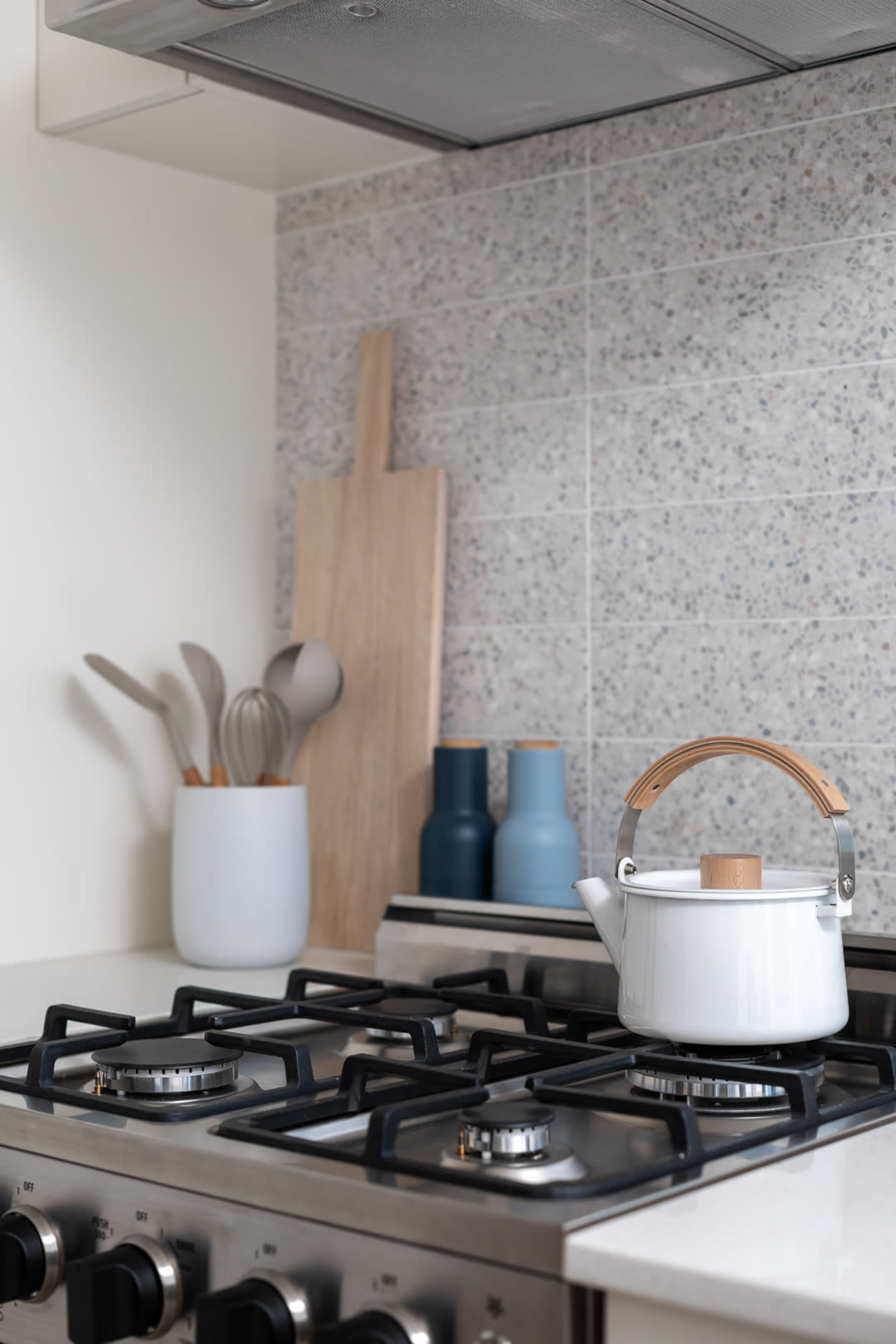 A white kettle with a wooden handle sits on a gas stove beneath a patterned backsplash, alongside a wooden cutting board and colored containers holding utensils.