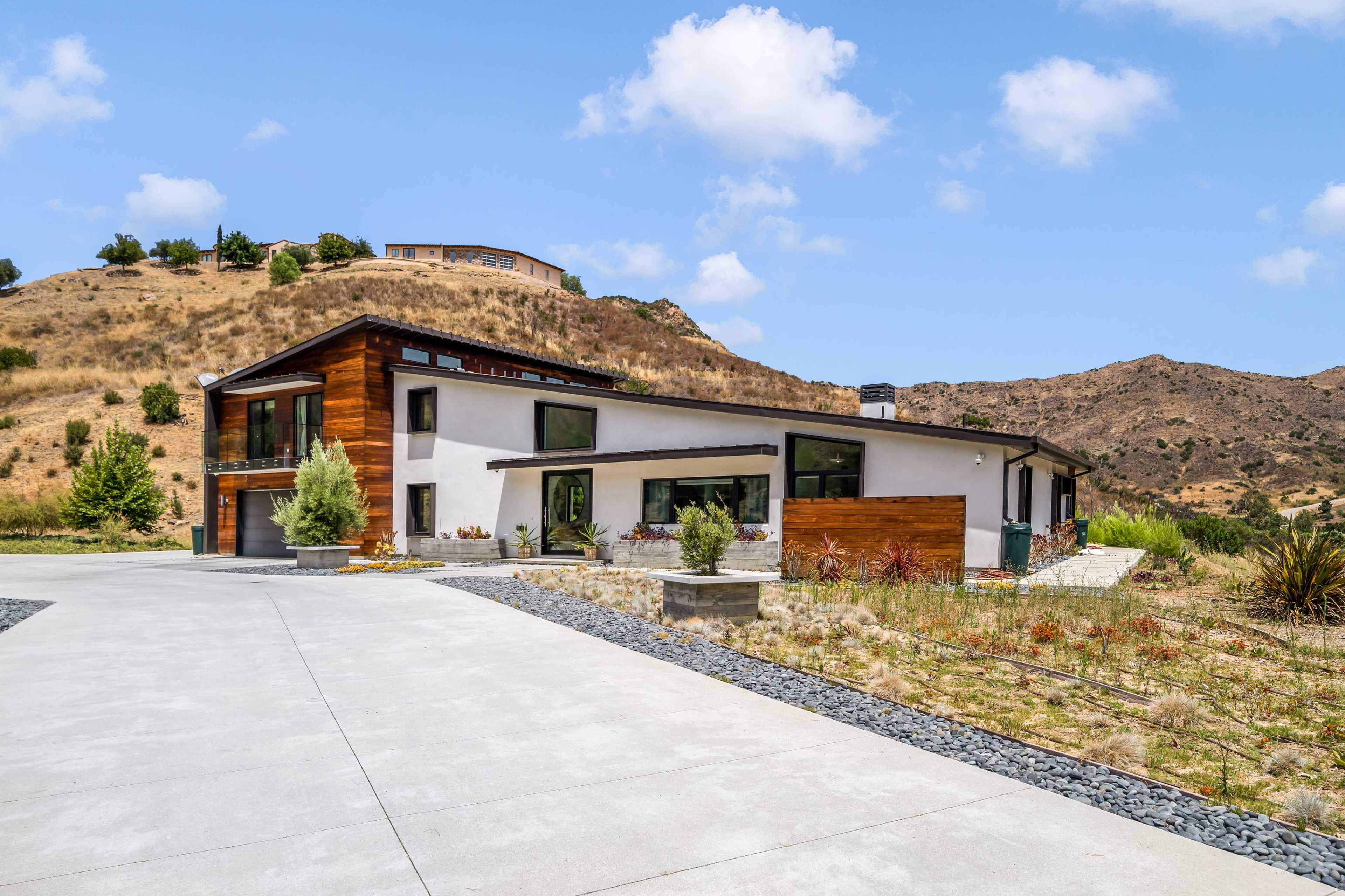 A modern house with a mix of wood and stucco exteriors, set against a hilly landscape with sparse vegetation.