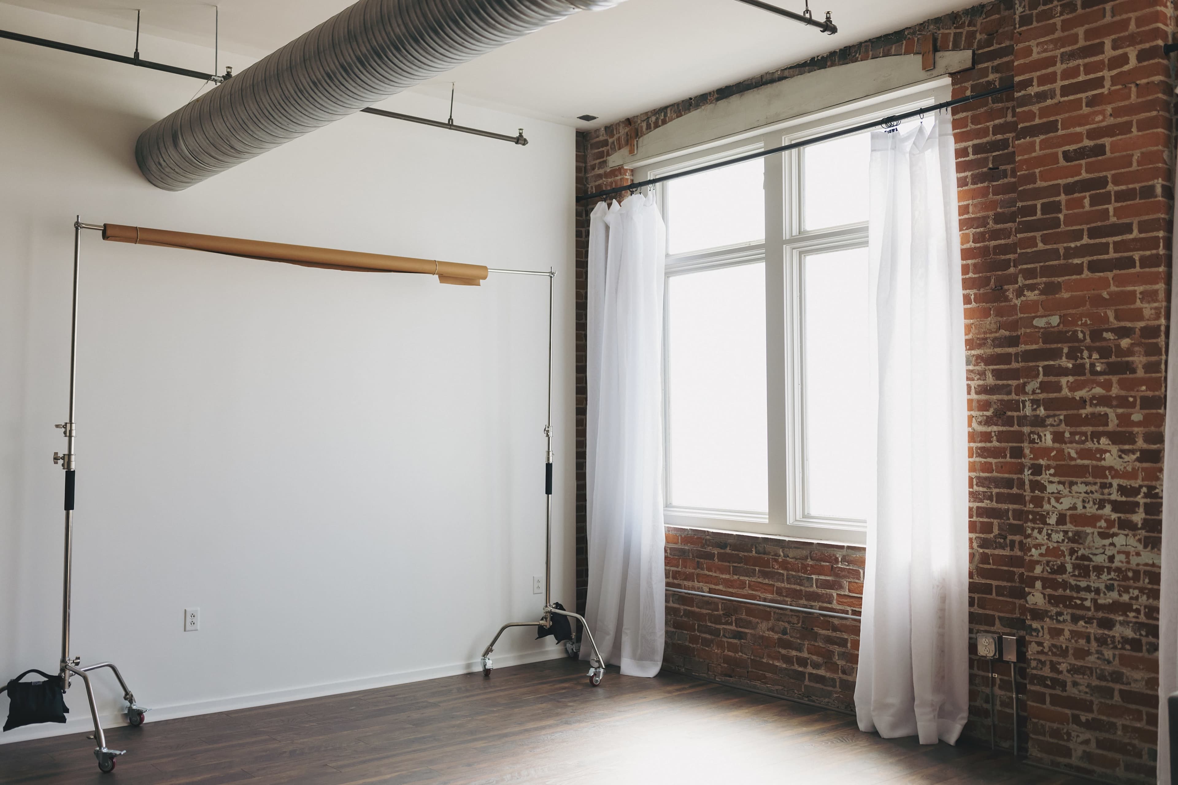 A minimalistic room with exposed brick walls, a large window dressed in white curtains, and a backdrop stand in the corner.