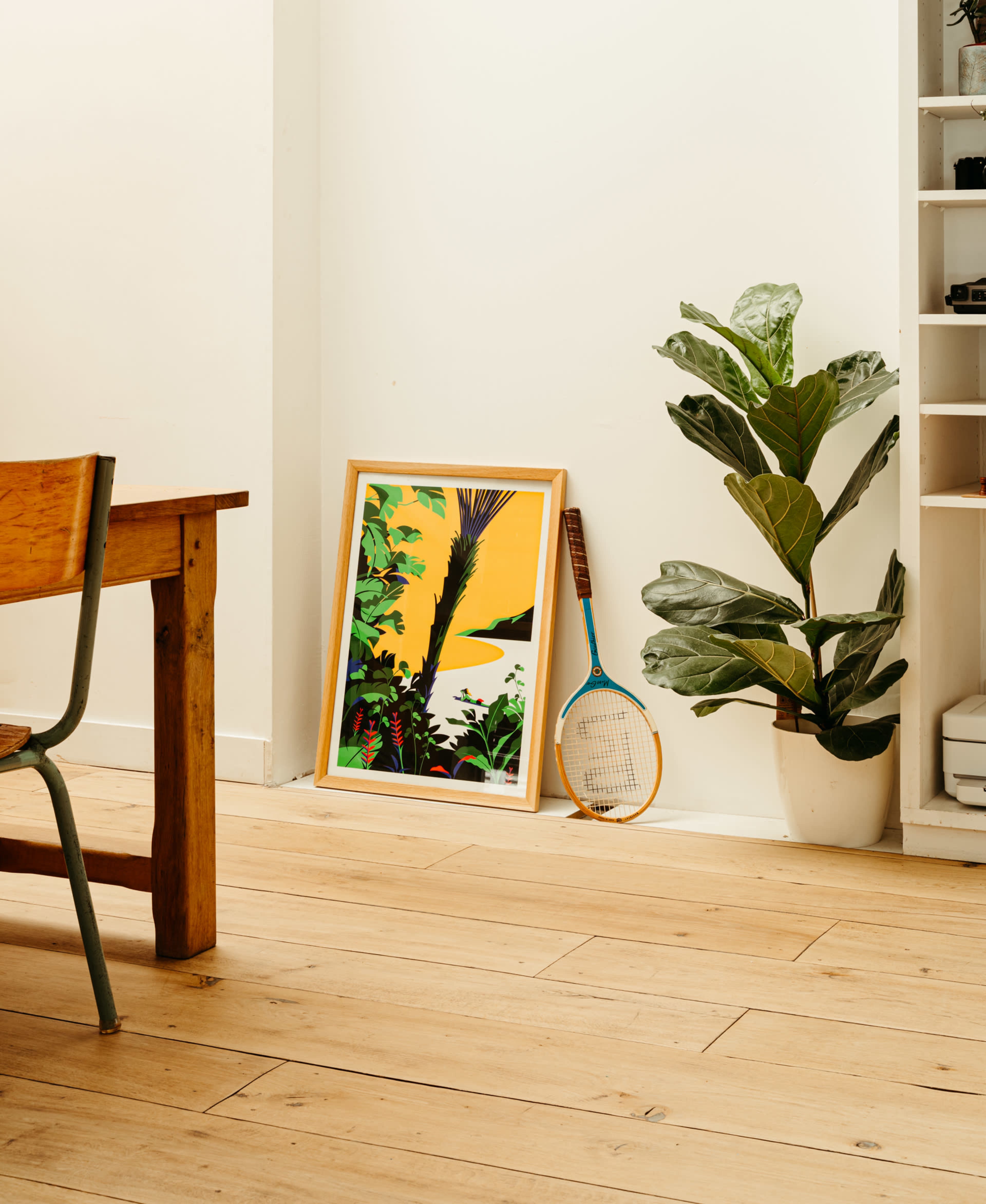 A framed artwork featuring green foliage leans against a wall next to a potted plant and a tennis racket in a room with wooden flooring and a wooden table.