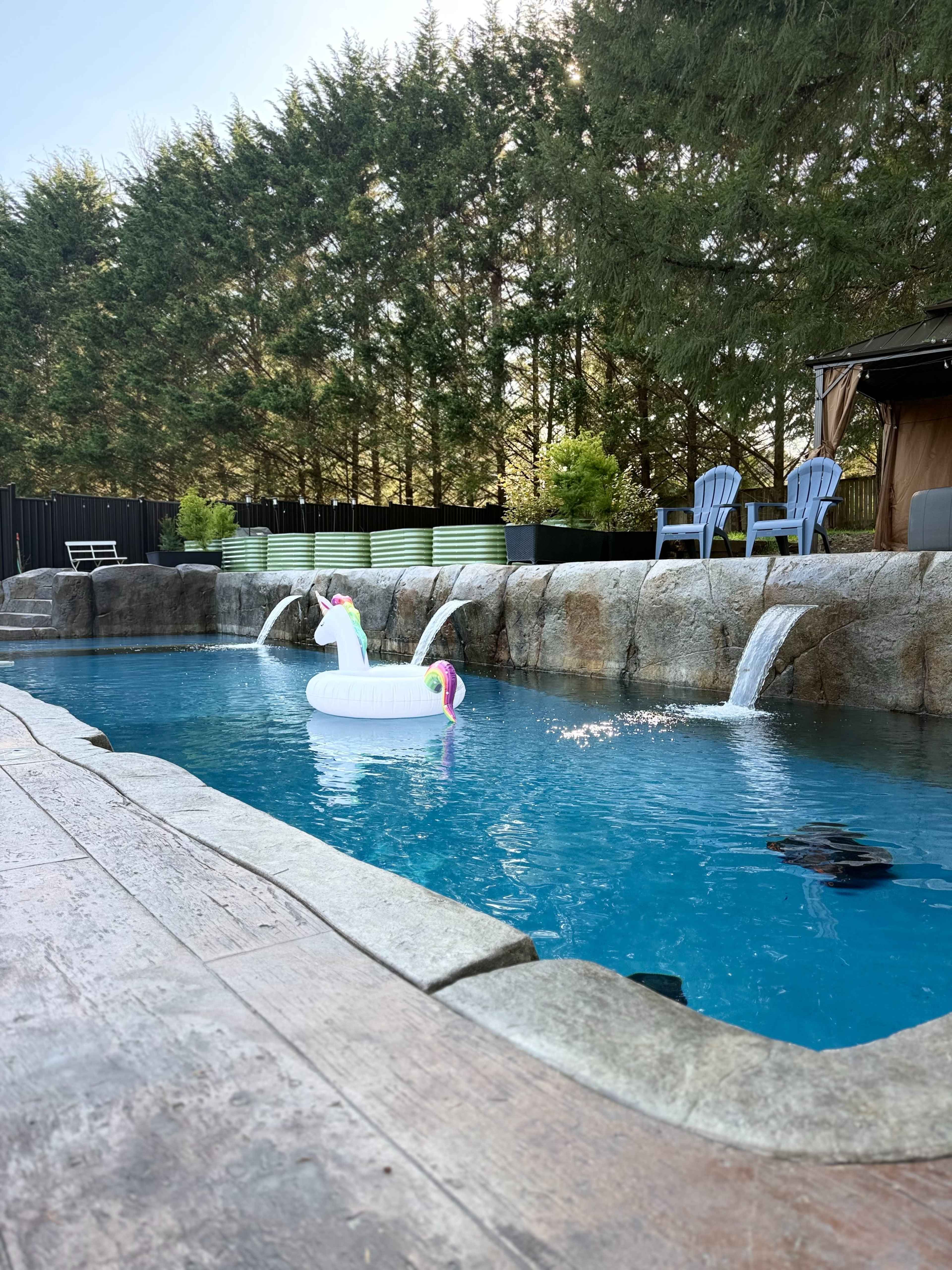 A unicorn float is positioned in a swimming pool surrounded by a stone edge and waterfalls, with lounge chairs in the background.