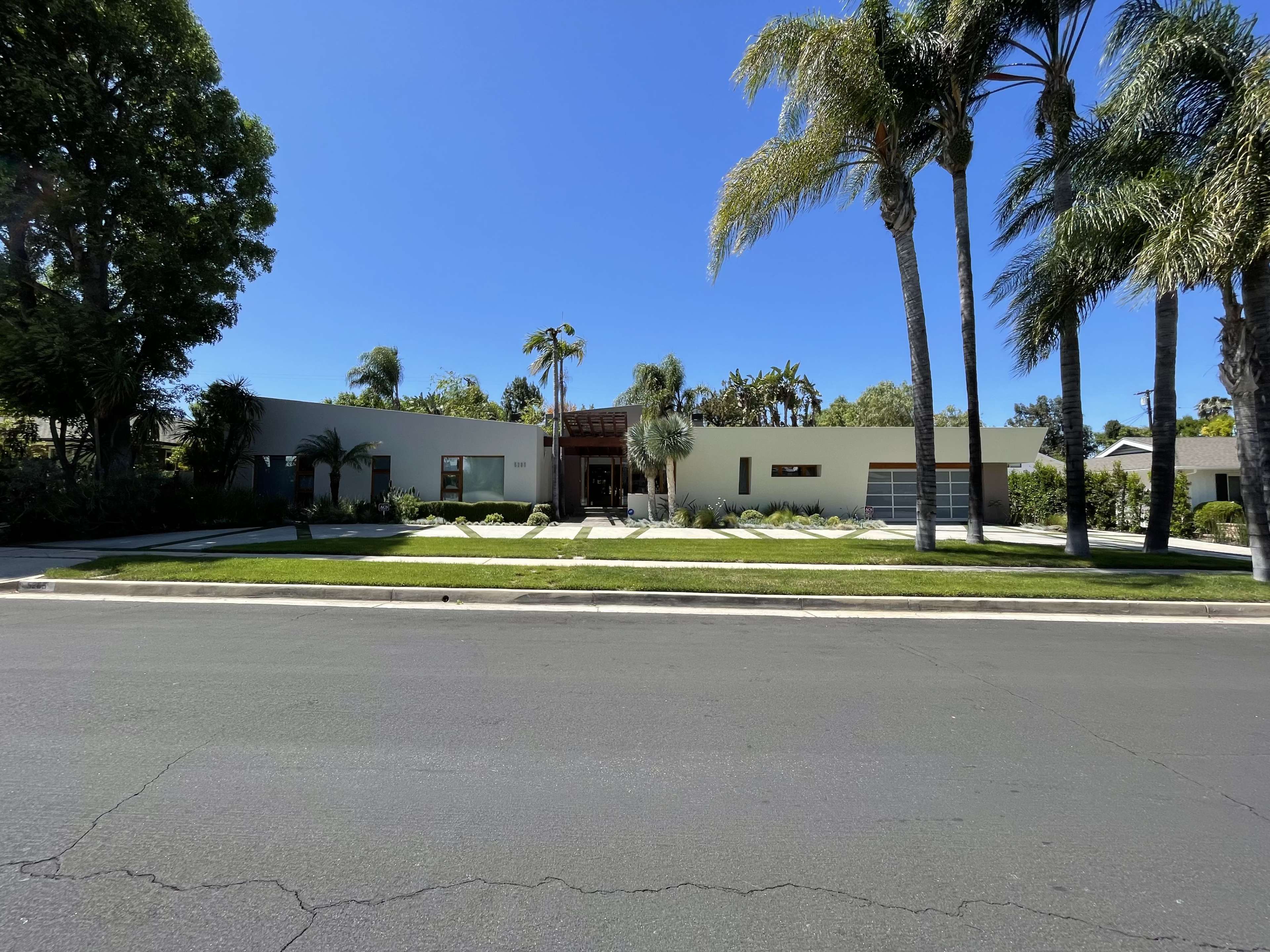 A modern single-story house with a flat roof and large windows is set behind a neatly manicured lawn and palm trees.