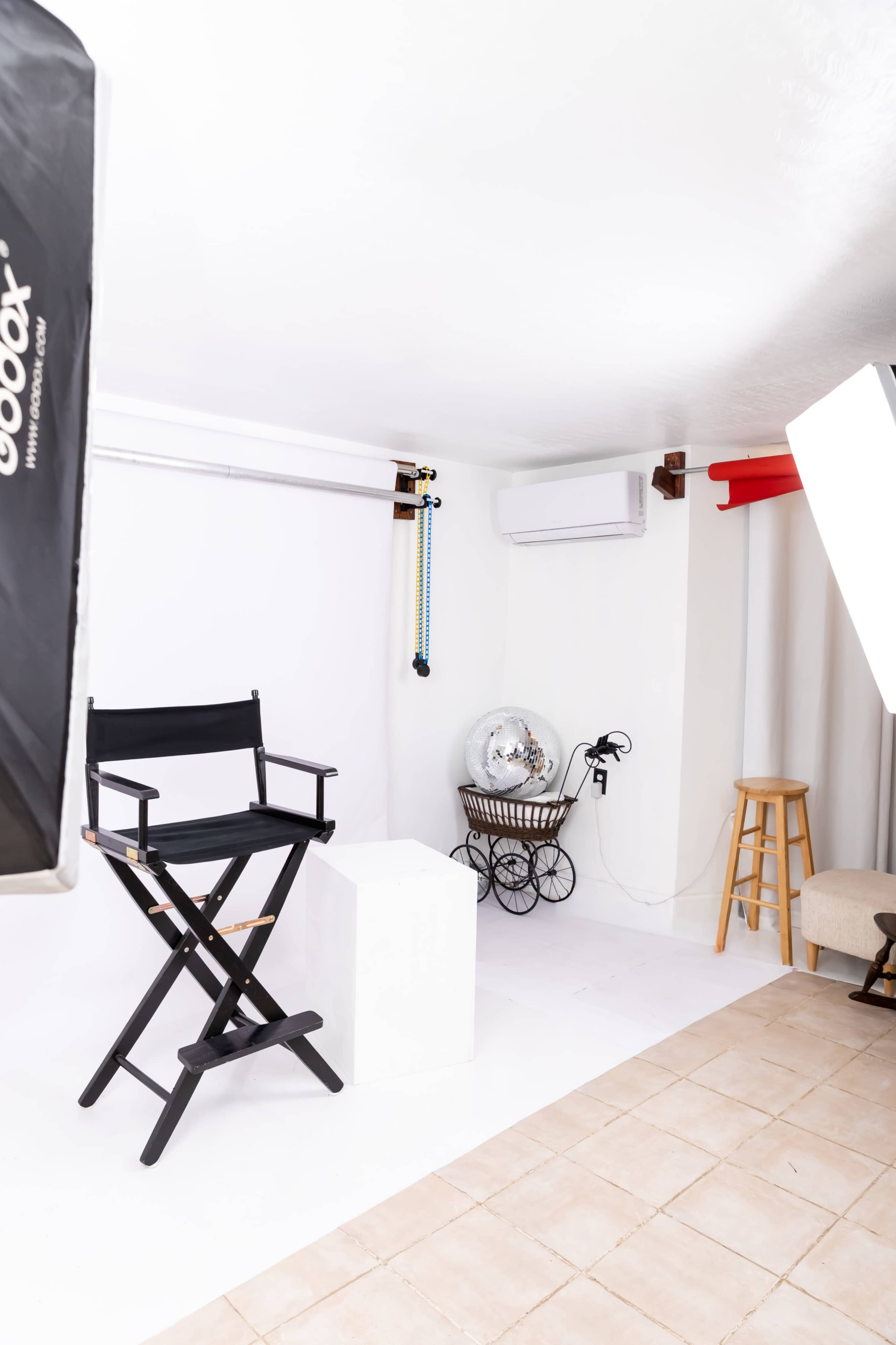 The image shows a photography studio with a black director's chair, a white backdrop, lighting equipment, and various props set up in a well-lit room.