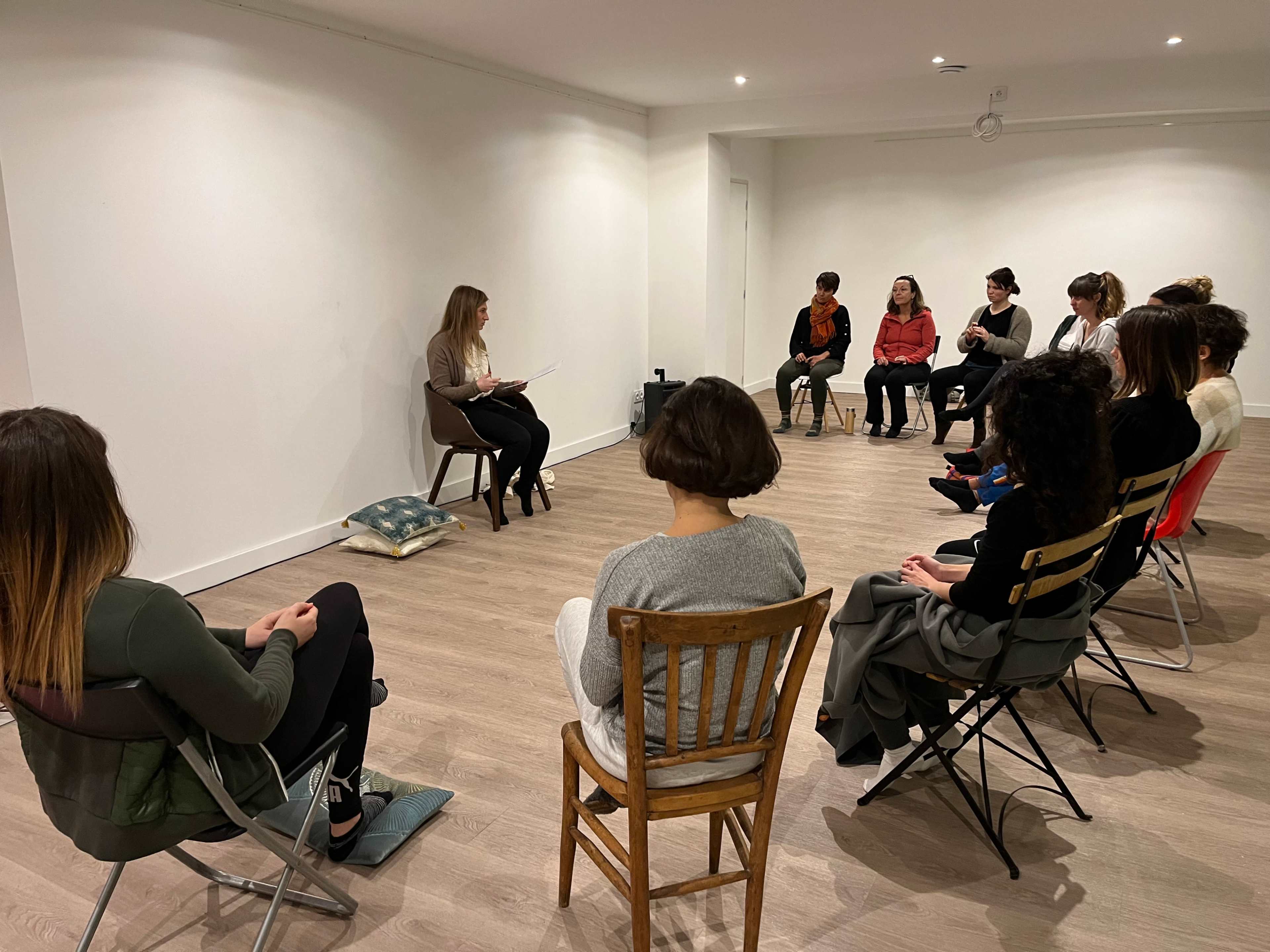 A group of ten people sits in a circle facing a speaker who is sitting on a chair and reading from a paper.
